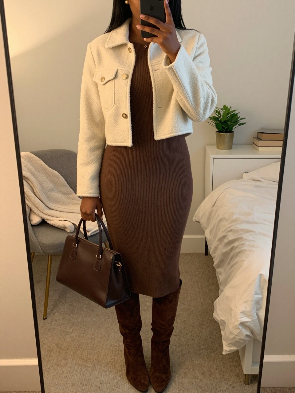 Mirror selfie of a Black woman wearing a fitted mocha ribbed-knit midi dress, chocolate-brown suede knee-high boots, a cropped cream bouclé jacket, a structured dark brown handbag, and small gold stud earrings, clearly showing the exact clothing items, colors, fabrics, and accessories listed, holding a phone in front of a cozy bedroom mirror setup, cropped at the neck so the face does not show, relaxed casual selfie pose, evening golden-hour lighting, a folded blanket on a chair and a small plant on a dresser, cozy and realistic, latest iPhone photo quality.