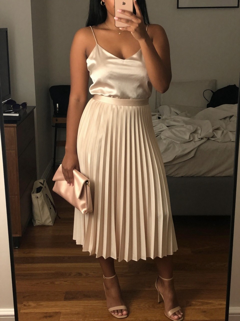Image prompt mirror selfie of a black curvy woman wearing a champagne pleated midi skirt with an ivory satin cami top, nude heeled sandals, a blush satin mini clutch, and small gold hoop earrings, holding a phone in front of a mirror, cropped at NECK so face doesn't show, with a natural casual selfie pose, in a city apartment, warm moody lighting, including small lived-in details like a tote bag on the floor, sunglasses on a dresser, or soft bedding in the background, latest iPhone photo quality.
