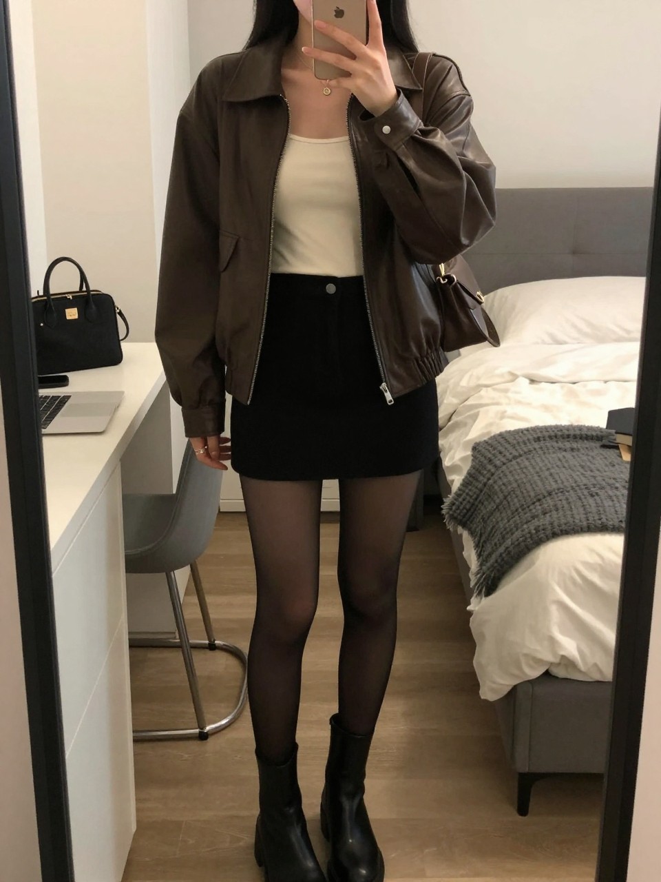 Mirror selfie of a Latina woman wearing a chocolate brown leather jacket, fitted cream tank top, black high-waisted mini skirt, black sheer tights, black ankle boots, and a brown shoulder bag, holding a phone in front of a mirror, cropped at NECK so the face does not show, relaxed casual selfie pose, in a stylish city apartment, warm moody lighting, with a laptop on the bed and a cosmetic bag on the counter, latest iPhone photo quality.