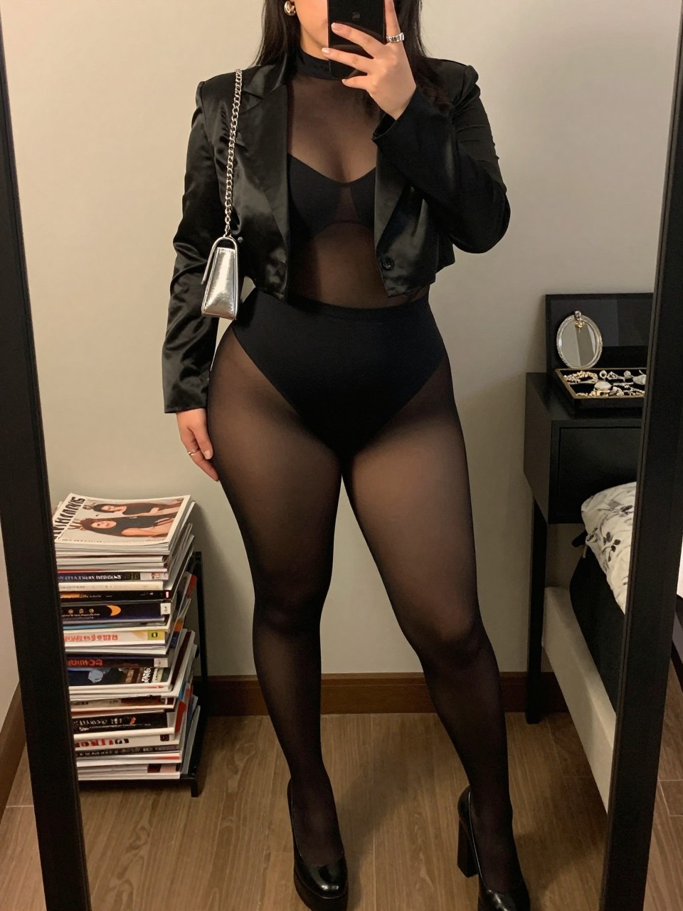 Mirror selfie of a plus size woman wearing a semi-sheer black mesh catsuit with long sleeves layered over a fitted opaque black bodysuit, a cropped black satin blazer worn open, black platform pumps, a small silver chain-strap bag, and silver cuff earrings, clearly showing the exact clothing layers, mesh texture, black satin, footwear, and accessories listed, holding a phone in front of a mirror, cropped at the neck so her face is hidden, relaxed bold selfie pose, in a cozy bedroom mirror setup with warm moody lighting, a stack of fashion magazines on a side table and a jewelry tray on the vanity, realistic edgy nightclub appearance, latest iPhone photo quality.