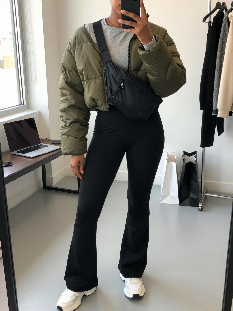 Mirror selfie of a Black woman wearing black flared leggings, a fitted heather-grey long-sleeve top, a cropped olive-green quilted puffer jacket, chunky white sneakers, and a black nylon belt bag worn across the chest, clearly showing the exact clothing items, colors, fabrics, and accessories listed, holding a phone in front of a fashion studio mirror corner, cropped at the neck so the face does not show, relaxed casual selfie pose, natural window light, laptop on a bench and shopping bags beside a chair, believable casual setting, latest iPhone photo quality.