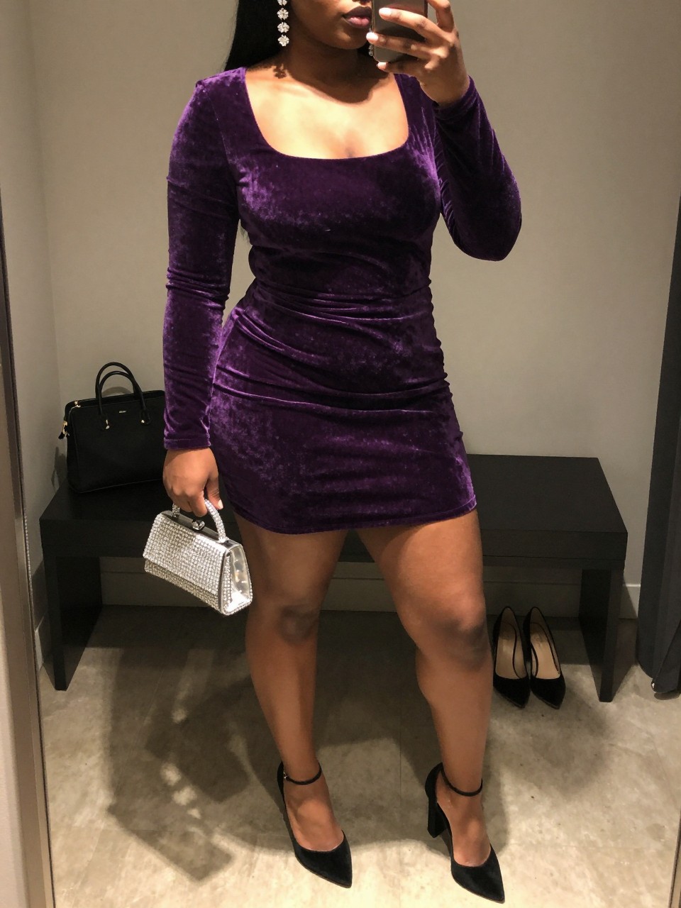 Mirror selfie of a black plus size woman wearing a plum purple velvet mini dress with long fitted sleeves, a deep square neckline, and soft gathering at the waist, black velvet block heels, a clear crystal-covered clutch, and crystal drop earrings, clearly showing the exact clothing items, plum velvet, gathering, colors, footwear, and accessories listed, holding a phone in front of a mirror, cropped at the neck so her face does not show, relaxed elegant selfie pose, in a minimal dressing room with warm moody lighting, a cosmetic bag on the counter and a pair of heels on the floor nearby, realistic upscale nightclub outfit, latest iPhone photo quality.