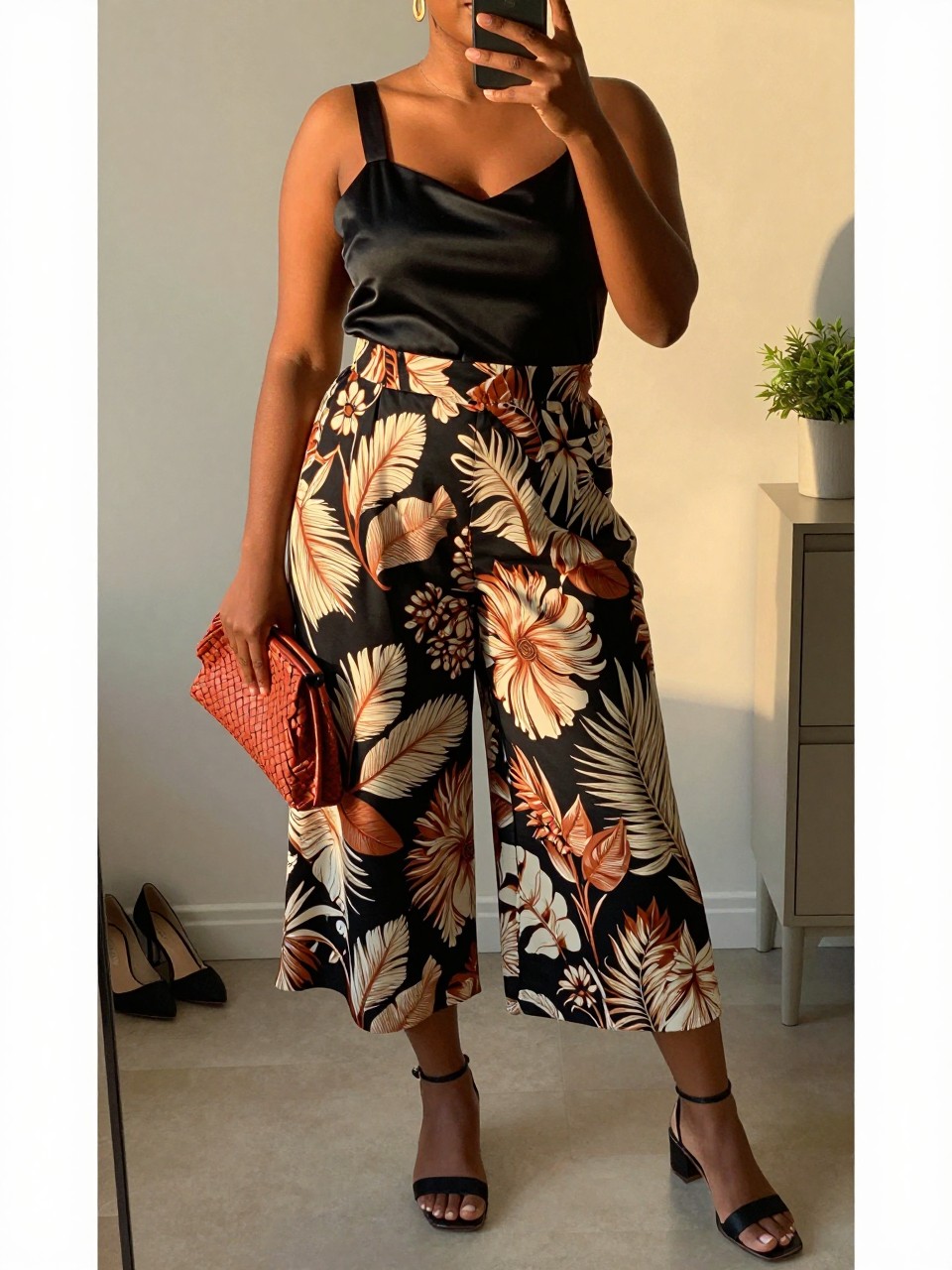 Mirror selfie of a black plus size woman wearing black, cream, and terracotta botanical-print palazzo shorts with a high waist and wide flowing legs, a fitted black satin camisole with wide supportive straps, black flat ankle-strap sandals, a terracotta woven clutch, and gold drop earrings, clearly showing the exact clothing items, botanical print, colors, satin texture, footwear, and accessories listed, holding a phone in front of a mirror, cropped at the neck so her face does not show, relaxed elegant selfie stance, in a minimal dressing room with evening golden-hour lighting, a pair of heels on the floor nearby and a small plant on a dresser, realistic summer evening outfit, latest iPhone photo quality.