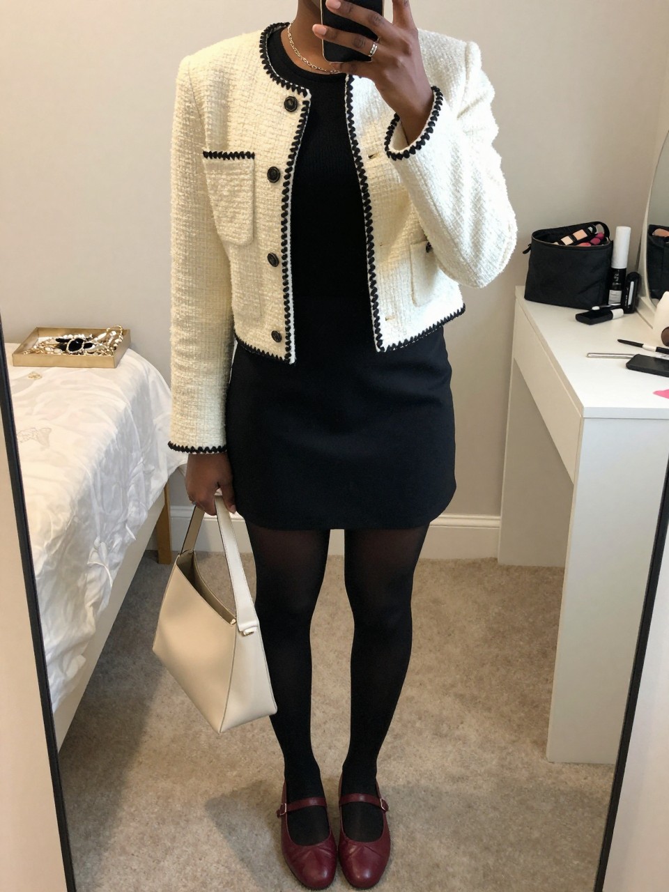 Mirror selfie of a Black woman wearing a cropped cream tweed jacket with black trim, fitted black crew-neck top, black A-line mini skirt, sheer black tights, burgundy leather ballet flats, and a structured cream shoulder bag, clearly showing the exact clothing items, colors, fabrics, and accessories listed, holding a phone in front of a feminine-themed bedroom mirror, cropped at the neck so the face does not show, relaxed casual selfie pose, soft indoor lighting, a jewelry tray on the vanity and a makeup bag on the counter, polished but natural, latest iPhone photo quality.