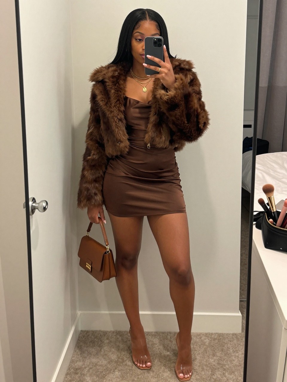 mirror selfie of a Black woman wearing the exact outfit described above, clearly showing a chocolate brown satin slip mini dress, brown cropped faux fur jacket, clear strap heels, gold layered necklace, and brown mini handbag, holding a phone in front of a mirror, cropped at NECK so the face does not show, with a confident casual selfie pose, in a feminine-themed bedroom, soft indoor lighting, including a handbag hanging on a door handle and a makeup brush or cosmetic bag on the counter, latest iPhone photo quality.