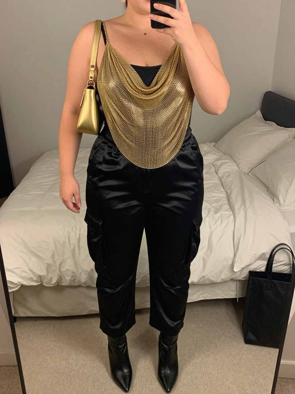 Mirror selfie of a plus size woman wearing a gold chainmail-style draped top layered over a fitted black bandeau, high-waisted black satin cargo pants with flat side pockets, black pointed ankle boots, a small gold leather shoulder bag, and black stud earrings, clearly showing the exact clothing layers, gold chain texture, black satin, pockets, footwear, and accessories listed, holding a phone in front of a mirror, cropped at the neck so the face does not show, relaxed glamorous selfie stance, in a feminine-themed bedroom with soft indoor lighting, soft pillows on an unmade bed and a tote bag resting on the floor, realistic fashion-forward clubwear appearance, latest iPhone photo quality.
