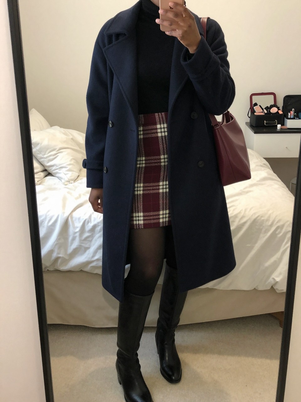 Mirror selfie of a Black woman wearing a burgundy, charcoal, and cream plaid wool mini skirt, a fitted black turtleneck, an oversized navy wool peacoat, opaque black tights, black leather knee-high boots, and a burgundy shoulder bag, clearly showing the exact clothing items, colors, fabrics, and accessories listed, holding a phone in front of a feminine-themed bedroom mirror, cropped at the neck so the face does not show, relaxed casual selfie pose, soft indoor lighting, soft pillows on an unmade bed and a makeup bag on the counter, natural lived-in environment, latest iPhone photo quality.