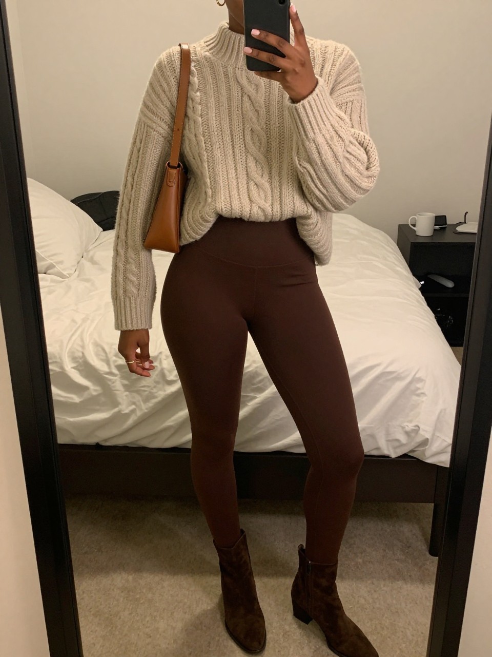 Mirror selfie of a Black woman wearing espresso-brown high-waisted leggings, an oversized oatmeal cable-knit tunic sweater, dark brown suede ankle boots, a tan leather shoulder bag, and small gold hoop earrings, clearly showing the exact clothing items, colors, fabrics, and accessories listed, holding a phone in front of a cozy bedroom mirror setup, cropped at the neck so the face does not show, relaxed casual selfie pose, warm moody lighting, soft pillows on an unmade bed and a coffee mug on a bedside table, realistic lived-in setting, latest iPhone photo quality.