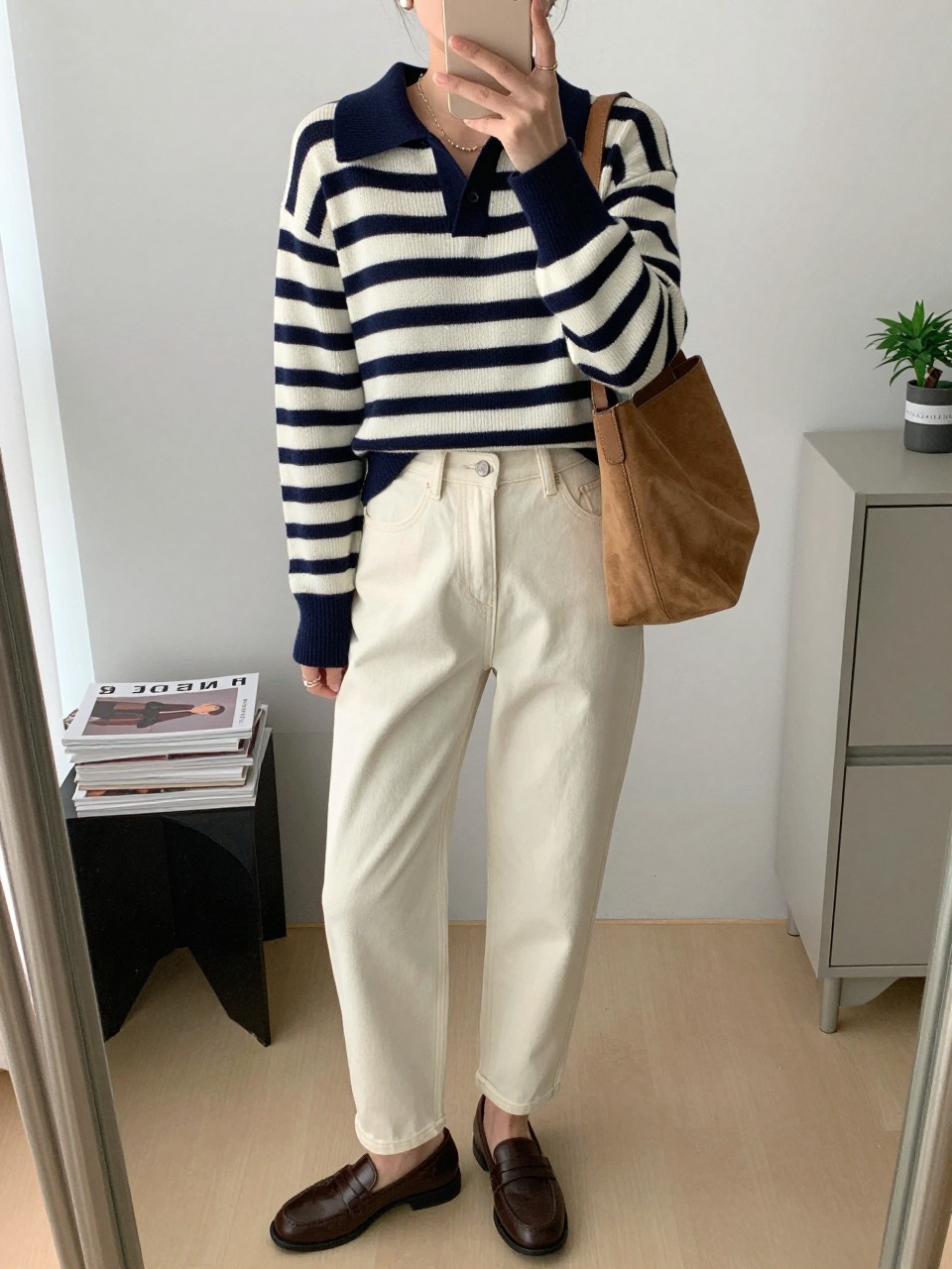 Mirror selfie of a woman wearing a fitted navy and cream striped polo-knit sweater, high-waisted cream barrel-leg jeans, dark brown leather loafers, a tan suede shoulder bag, and small pearl stud earrings, clearly showing the exact clothing items, colors, fabrics, and accessories listed, holding a phone in front of a minimal dressing room mirror, cropped at the neck so the face does not show, relaxed casual selfie pose, natural window light, a stack of fashion magazines on a side table and a small plant on a dresser, realistic and uncluttered, latest iPhone photo quality.