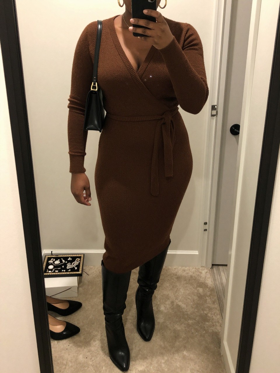 Mirror selfie of a curvy Black woman wearing a chocolate brown wrap sweater midi dress, black knee-high leather boots, gold hoop earrings, and a small black shoulder bag, holding a phone in front of a mirror, cropped at NECK so the face does not show, relaxed casual selfie pose, in a walk-in closet mirror, warm indoor lighting, with a jewelry tray on the vanity and a pair of heels on the floor nearby, latest iPhone photo quality.