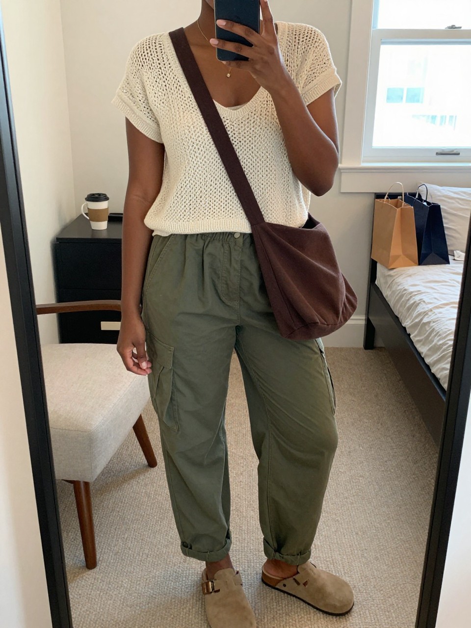 Mirror selfie of a Black woman wearing a cream short-sleeve crochet-knit top over a matching cream camisole, loose olive-green utility pants, tan suede clogs, and a chocolate-brown canvas crossbody bag, clearly showing the exact clothing items, colors, fabrics, and accessories listed, holding a phone in front of a cozy bedroom mirror setup, cropped at the neck so the face does not show, relaxed casual selfie pose, natural window light, a coffee mug on a bedside table and shopping bags beside a chair, casual realistic atmosphere, latest iPhone photo quality.