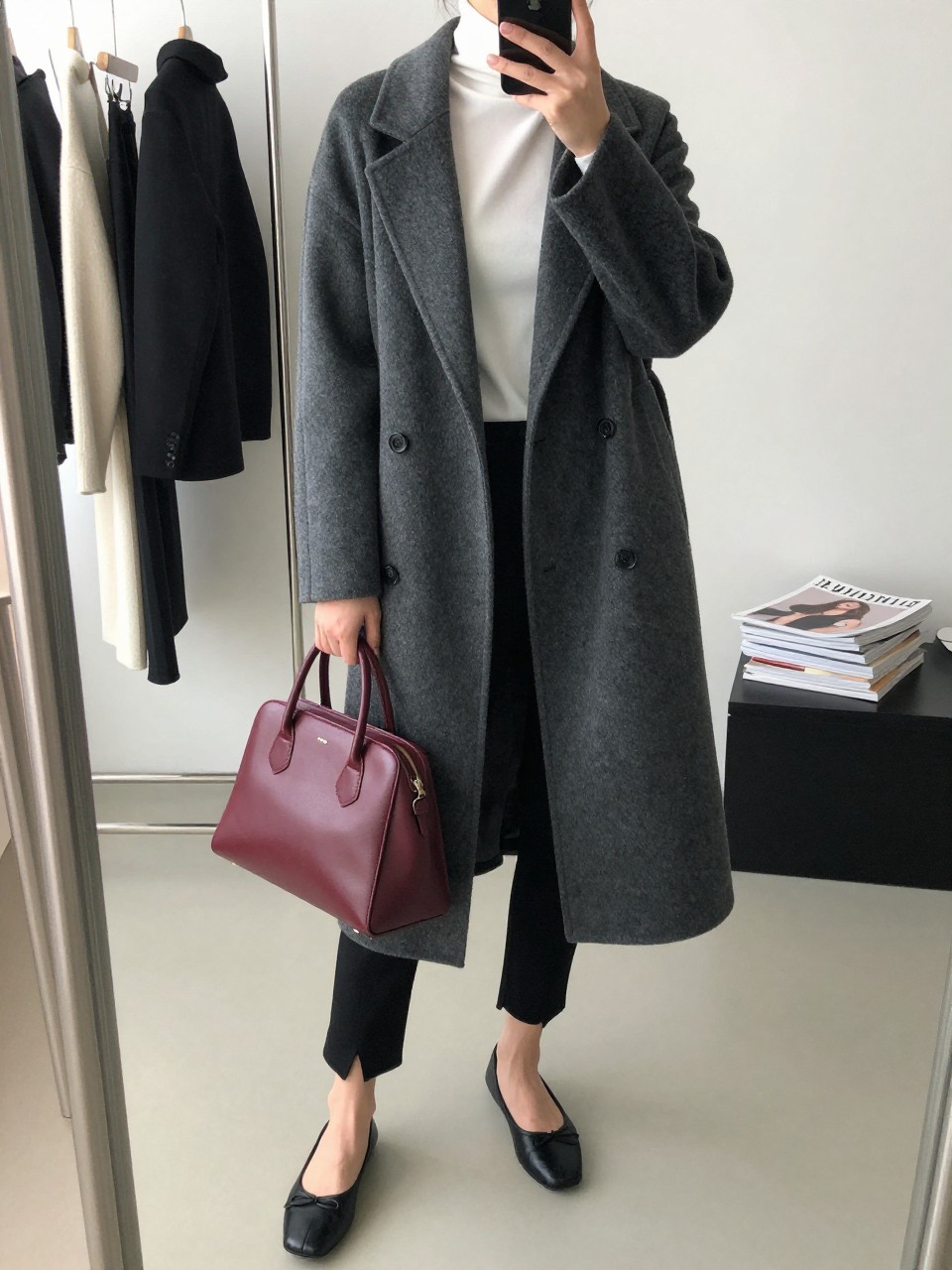 Mirror selfie of a woman wearing black split-hem leggings, a fitted white turtleneck, a long charcoal-grey wool coat, black leather ballet flats with square toes, and a structured oxblood handbag, clearly showing the exact clothing items, colors, fabrics, and accessories listed, holding a phone in front of a minimal dressing room mirror, cropped at the neck so the face does not show, relaxed casual selfie pose, natural window light, clothing rack with garments behind and a stack of fashion magazines on a side table, polished realistic scene, latest iPhone photo quality.