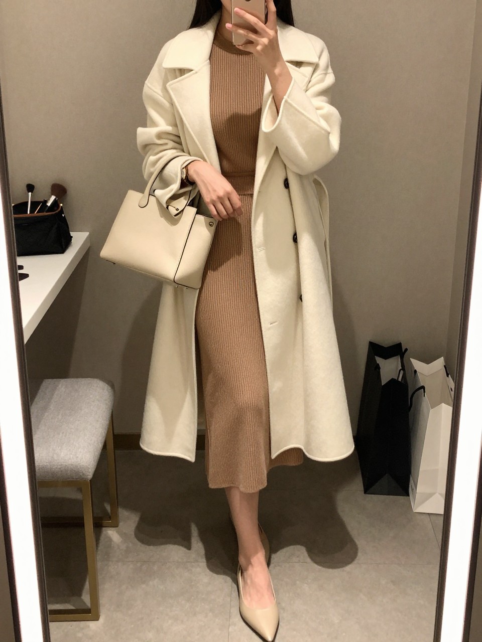 mirror selfie of a woman wearing the exact outfit described above, clearly showing a mocha knit top, matching mocha knit midi skirt, long ivory coat, beige pointed flats, a cream shoulder bag, and a gold watch, holding a phone in front of a mirror, cropped at NECK so the face does not show, with a relaxed casual selfie pose, in a minimal dressing room, warm moody lighting, with a makeup brush or cosmetic bag on the counter and shopping bags beside a chair, latest iPhone photo quality.