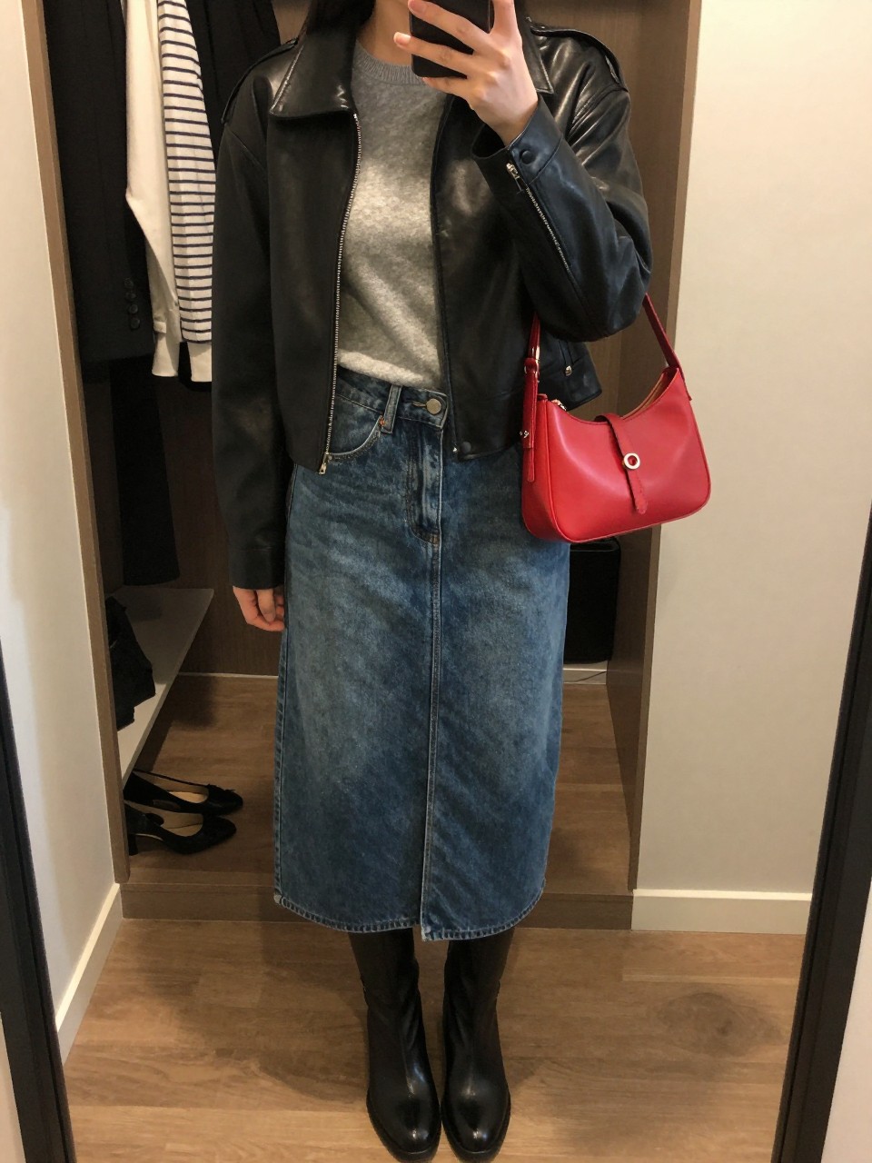 Mirror selfie of a woman wearing a cropped black leather jacket, fitted soft-grey crew-neck knit top, medium-wash denim midi skirt with a front slit, black leather knee-high boots, and a vivid red mini shoulder bag, clearly showing the exact clothing items, colors, fabrics, and accessories listed, holding a phone in front of a walk-in closet mirror, cropped at the neck so the face does not show, relaxed casual selfie pose, warm moody lighting, an open closet with clothes visible and a pair of heels on the floor nearby, natural and unstaged, latest iPhone photo quality.