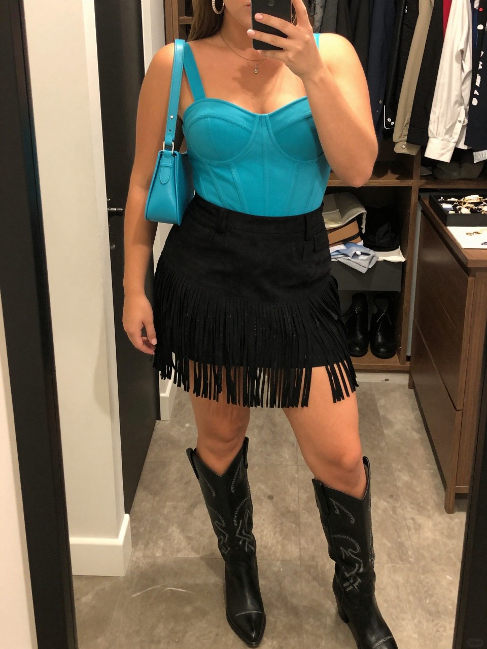 Mirror selfie of a plus size woman wearing a high-waisted black suede mini skirt with layered fringe, a structured turquoise corset-style top with wide supportive straps, black leather knee-high cowboy boots with silver stitching, a small turquoise leather shoulder bag, and chunky silver hoop earrings, clearly showing the exact clothing items, black suede fringe, turquoise top, stitched boots, colors, and accessories listed, holding a phone in front of a mirror, cropped at the neck so her face does not appear, relaxed powerful selfie stance, in a walk-in closet mirror with warm moody lighting, an open closet with clothes visible and a jewelry tray on the vanity, realistic vaquera night-out outfit, latest iPhone photo quality.