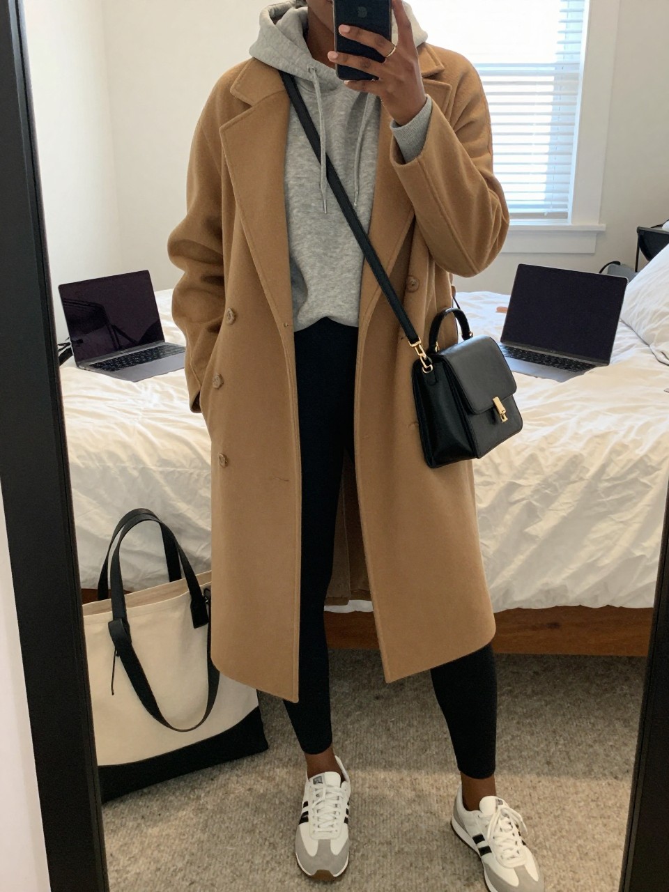 Mirror selfie of a Black woman wearing a soft heather-grey fitted hoodie beneath a knee-length camel wool wrap coat, matte black high-waisted leggings, white and grey retro sneakers, and a structured black crossbody bag, clearly showing the exact clothing items, colors, fabrics, and accessories listed, holding a phone in front of a cozy bedroom mirror setup, cropped at the neck so the face does not show, relaxed casual selfie pose, natural window light, a laptop on the bed and a tote bag resting on the floor, casual realistic setting, latest iPhone photo quality.