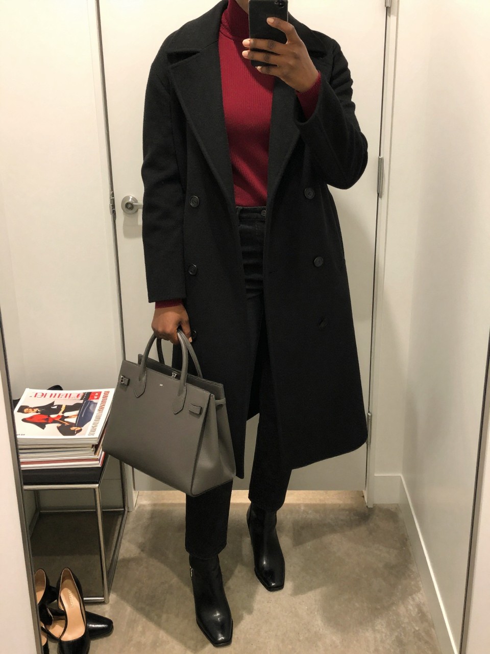 Mirror selfie of a Black woman wearing a long black wool coat, fitted cherry-red mock-neck sweater, black straight-leg jeans, black leather square-toe ankle boots, and a structured charcoal-grey handbag, clearly showing the exact clothing items, colors, fabrics, and accessories listed, holding a phone in front of a minimal dressing room mirror, cropped at the neck so the face does not show, relaxed confident selfie pose, warm moody lighting, a stack of fashion magazines on a side table and a pair of heels on the floor nearby, sleek and realistic, latest iPhone photo quality.
