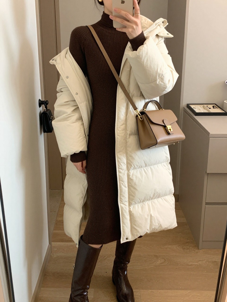 Mirror selfie of a woman wearing a fitted chocolate-brown ribbed midi sweater dress, a belted cream knee-length puffer coat, dark brown leather knee-high boots, a structured tan crossbody bag, and small gold stud earrings, clearly showing the exact clothing items, colors, fabrics, and accessories listed, holding a phone in front of a walk-in closet mirror, cropped at the neck so the face does not show, relaxed casual selfie pose, soft indoor lighting, a handbag hanging on a door handle and a jewelry tray on the vanity, warm natural setting, latest iPhone photo quality.