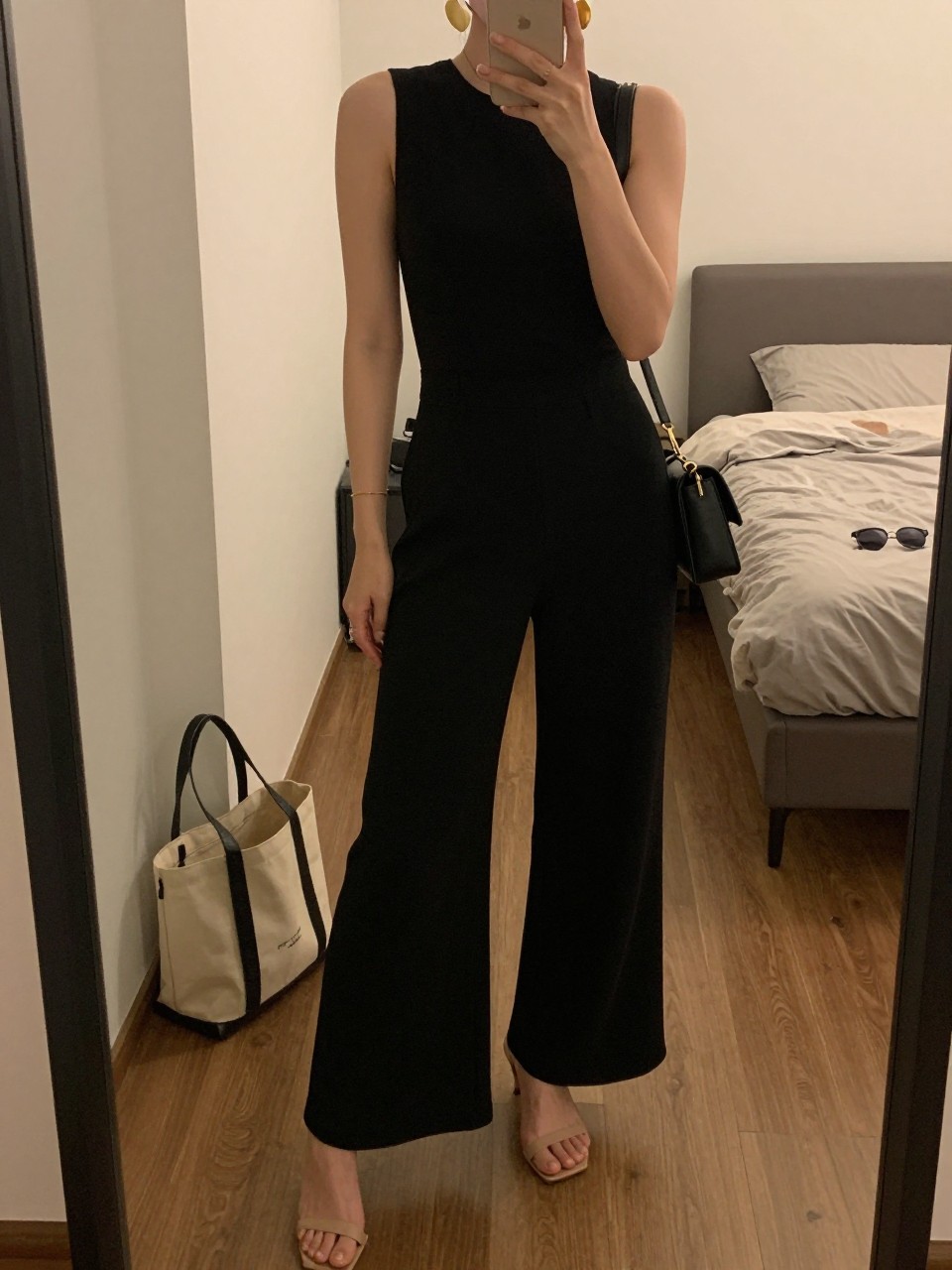 Image prompt mirror selfie of a woman wearing a black crepe wide-leg jumpsuit with nude heeled sandals, bold clean gold statement earrings, and a black mini shoulder bag, holding a phone in front of a mirror, cropped at NECK so face doesn't show, with a natural casual selfie pose, in a city apartment, warm moody lighting, including small lived-in details like a tote bag on the floor, sunglasses on a dresser, or soft bedding in the background, latest iPhone photo quality.