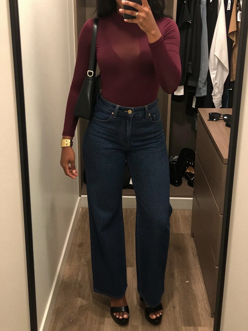 mirror selfie of a Black woman wearing the exact outfit described above, clearly showing a burgundy long-sleeve mesh bodysuit, dark blue high-waisted wide-leg jeans, black platform heels, black leather shoulder bag, and gold cuff bracelet, holding a phone in front of a mirror, cropped at NECK so the face does not show, with a confident casual selfie pose, in a walk-in closet mirror, warm moody lighting, including an open closet with clothes visible and sunglasses placed on a dresser, latest iPhone photo quality.