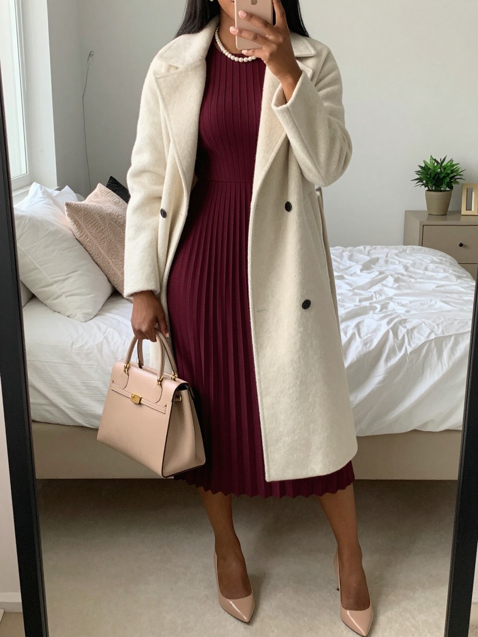 mirror selfie of a Black woman wearing the exact outfit described above, clearly showing a burgundy pleated midi dress, cream wool coat worn open, nude closed-toe pumps, a beige top-handle bag, and a pearl necklace, holding a phone in front of a mirror, cropped at NECK so the face does not show, with a relaxed casual selfie pose, in a feminine-themed bedroom, natural window light, with soft pillows on an unmade bed and a small plant on a dresser, latest iPhone photo quality.