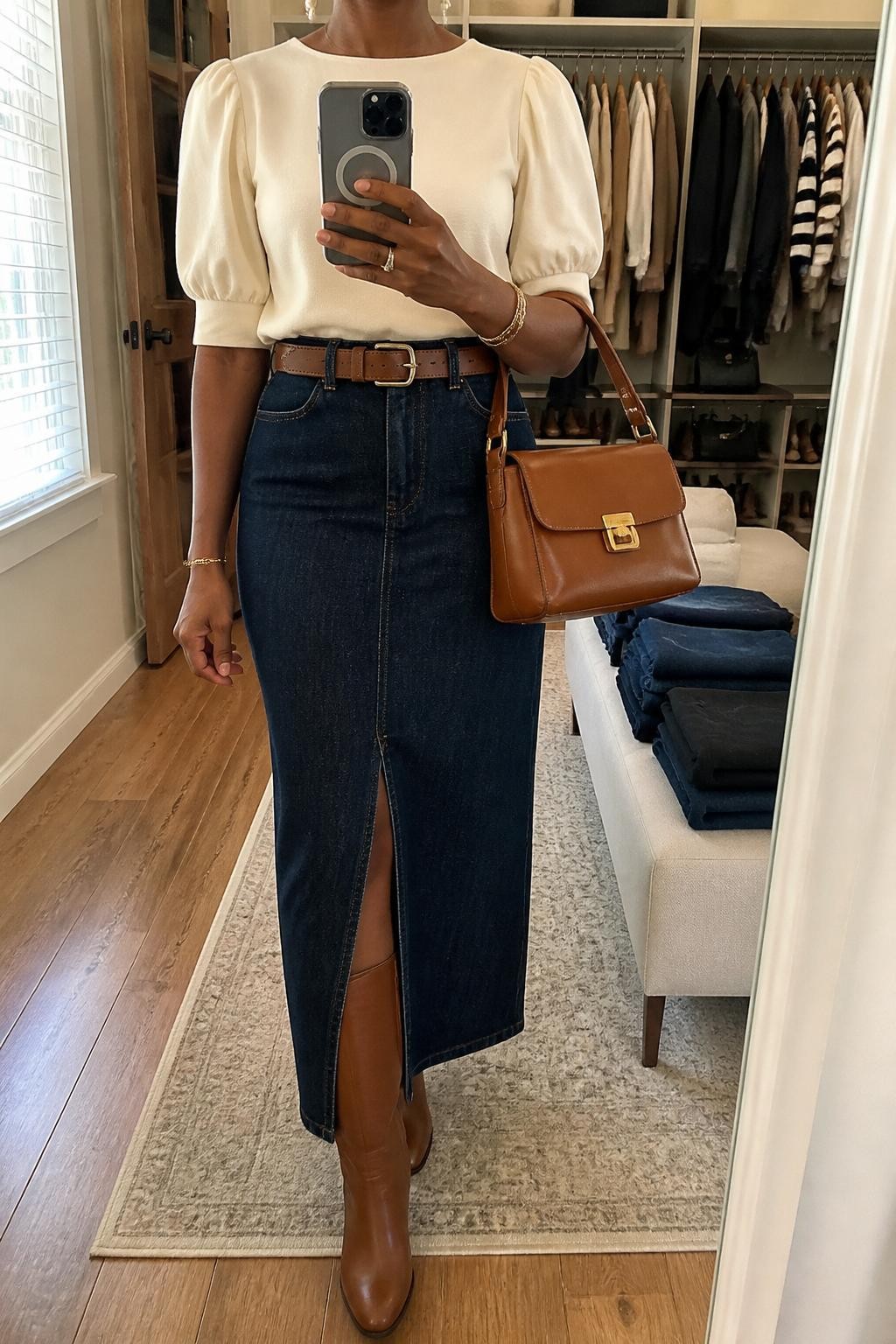 mirror selfie of a Black woman wearing the exact outfit described above, clearly showing a dark denim maxi skirt with a front slit, cream puff-sleeve blouse, cognac riding boots, tan waist belt, brown structured satchel, and pearl drop earrings, holding a phone in front of a mirror, cropped at NECK so the face does not show, with a relaxed casual selfie pose, in a walk-in closet mirror, natural window light, with open closet with clothes visible and folded denim jeans on a bench, latest iPhone photo quality.