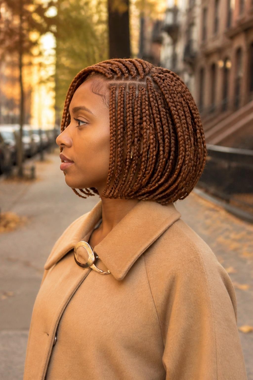 Image prompt: Photo of a Black woman wearing a chin-length copper box braid bob with a deep side part and slightly curved ends, side profile view, softly blurred city street background with scattered autumn leaves, golden hour glow, containing small gold earrings peeking through and a hair clip attached to her coat collar, realistic candid fall moment, latest iPhone photo quality.