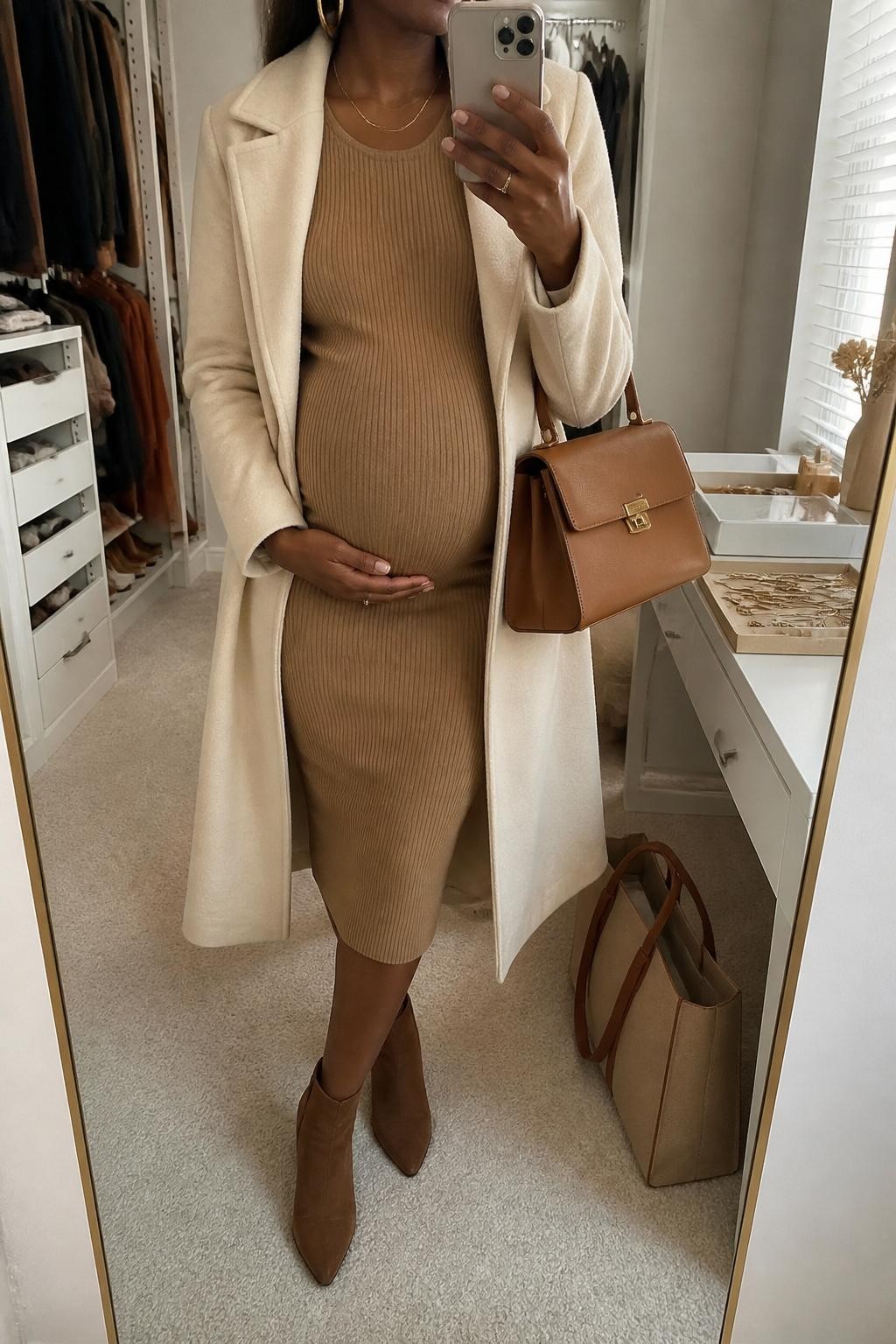 mirror selfie of a pregnant Black woman wearing a camel ribbed knit maternity midi dress, brown suede ankle boots, a cream wool coat, gold hoop earrings, and a tan structured handbag, clearly showing the same clothing items, colors, fabrics, and accessories listed in the Outfit Pieces section, holding a phone in front of a mirror, cropped at NECK so the face does not show, with a relaxed casual selfie pose, in a walk-in closet mirror, natural window light, including a tote bag resting on the floor and jewelry tray on the vanity, latest iPhone photo quality.