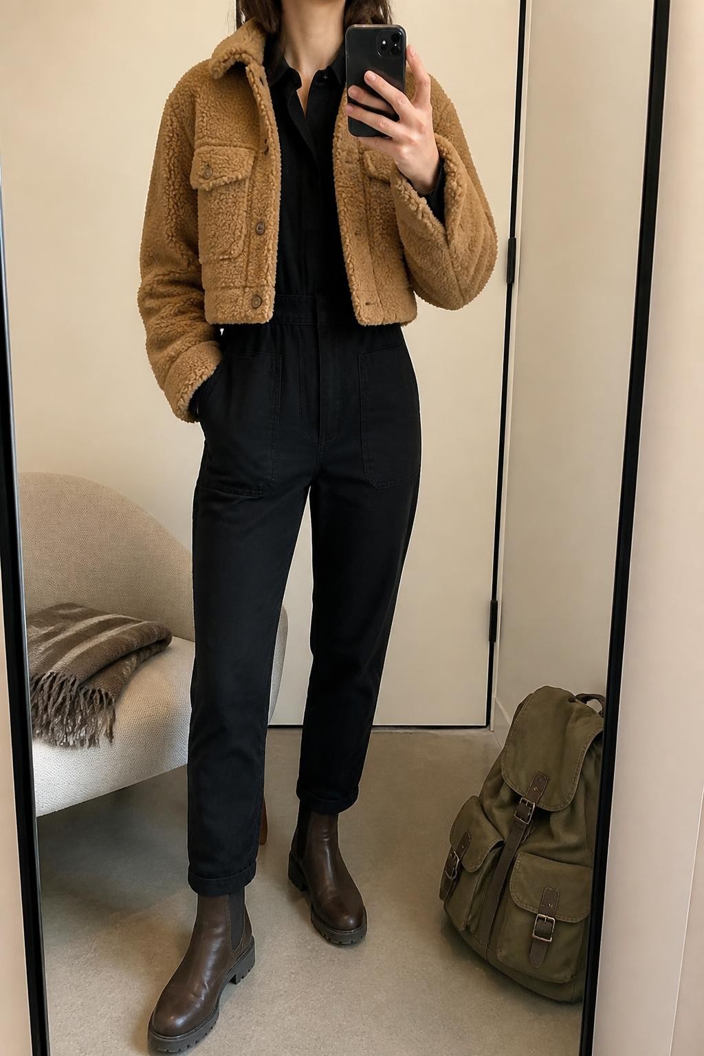 Mirror selfie of a woman wearing a black long-sleeve utility jumpsuit with a defined waist, cropped camel sherpa jacket, dark brown waterproof ankle boots, and a structured olive-green canvas backpack, clearly showing the exact clothing items, colors, fabrics, and accessories listed, holding a phone in front of a minimal dressing room mirror, cropped at the neck so the face does not show, relaxed casual selfie pose, soft indoor lighting, a backpack resting on the floor and a folded blanket on a chair, natural and uncluttered, latest iPhone photo quality.