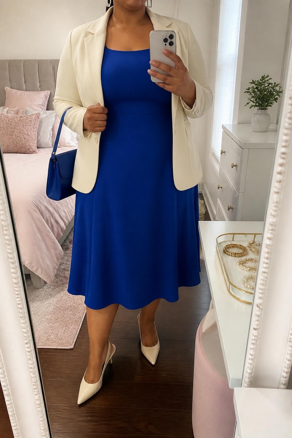 Mirror selfie of a plus size woman wearing a cobalt blue crepe A-line midi dress with short sleeves, a modest square neckline, and a softly fitted waist, a cream tailored blazer worn open, cream pointed slingback heels, a structured cobalt shoulder bag, and silver rectangular earrings, clearly showing the exact clothing items, cobalt dress, cream blazer, footwear, and accessories listed, holding a phone in front of a mirror, cropped at the neck so the face does not show, relaxed confident selfie pose, in a feminine-themed bedroom with natural window light, a jewelry tray on the vanity and a small plant on a dresser, realistic church celebration outfit, latest iPhone photo quality.