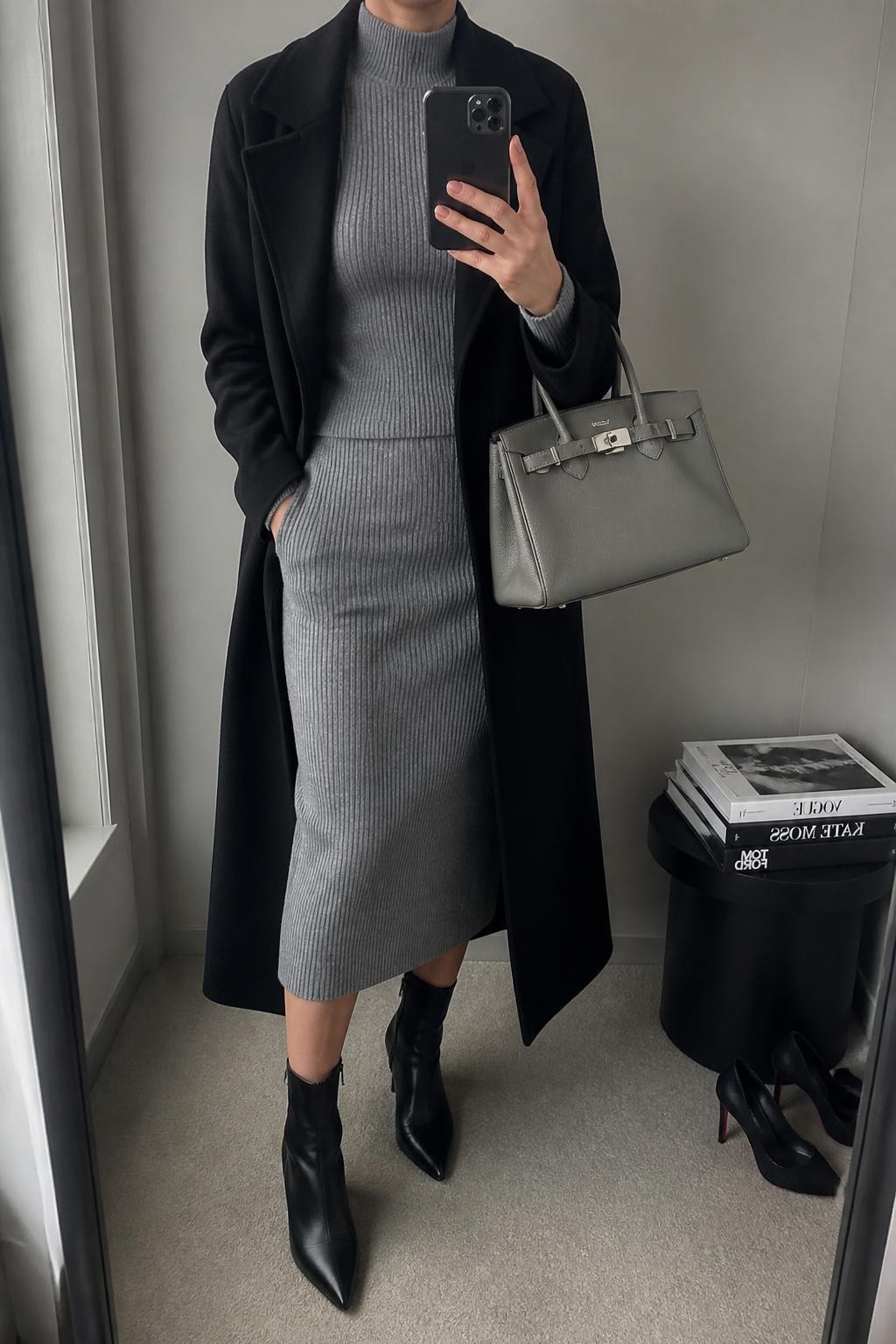 Mirror selfie of a woman wearing a heather-grey ribbed-knit midi skirt, a matching fitted grey mock-neck sweater, a long black wool coat, black leather pointed-toe ankle boots, and a structured silver-grey handbag, clearly showing the exact clothing items, colors, fabrics, and accessories listed, holding a phone in front of a minimal dressing room mirror, cropped at the neck so the face does not show, relaxed casual selfie pose, natural window light, a stack of fashion magazines on a side table and a pair of heels on the floor nearby, sleek realistic setting, latest iPhone photo quality.