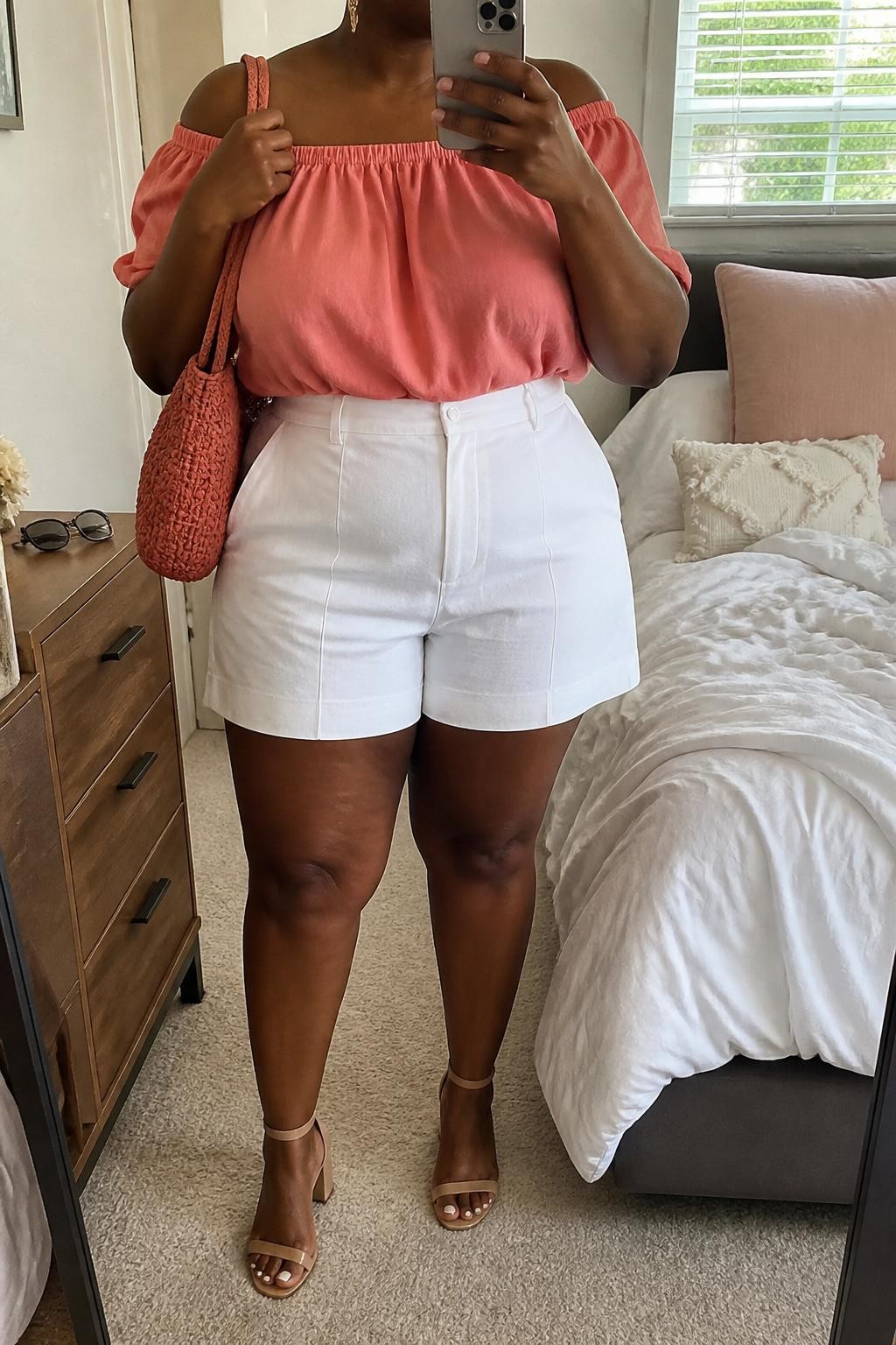 Mirror selfie of a plus size black woman wearing crisp white high-waisted tailored shorts with relaxed thigh openings, a coral pink off-shoulder blouse with short gathered sleeves, nude block-heel sandals, a coral woven shoulder bag, and delicate gold drop earrings, clearly showing the exact clothing items, white tailoring, coral blouse, gathered neckline, footwear, and accessories listed, holding a phone in front of a mirror, cropped at the neck so her face is hidden, relaxed cheerful selfie stance, in a cozy bedroom mirror setup with natural window light, sunglasses placed on a dresser and soft pillows on an unmade bed, realistic brunch-date outfit, latest iPhone photo quality.
