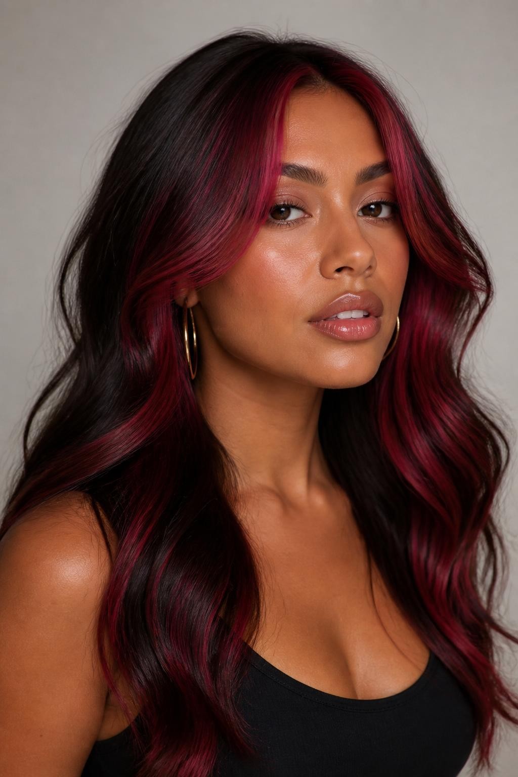 Image Prompt: A stylish woman with dark wavy hair and raspberry money piece highlights framing the face, rich dark brown base with bold raspberry-red front sections, loose waves with shine, warm medium-deep skin tone, glossy nude lips, subtle bronzed makeup, gold hoop earrings, fitted black top, clean neutral studio background, close-up beauty portrait, realistic DSLR photo, clear face-framing raspberry color detail, no text, (no watermarks on images)