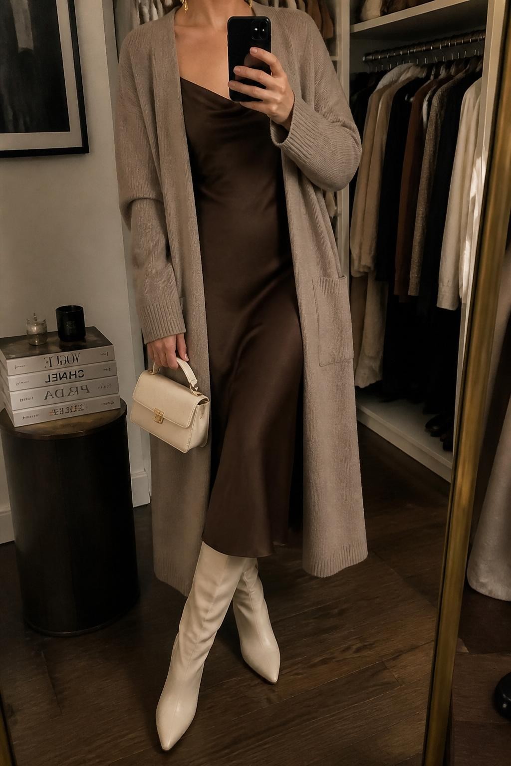 mirror selfie of a woman wearing the exact outfit described above, clearly showing a taupe long coatigan layered over a chocolate brown satin midi slip dress, cream knee-high boots, a cream mini handbag, and gold drop earrings, holding a phone in front of a mirror, cropped at NECK so the face does not show, with a relaxed casual selfie pose, in a walk-in closet mirror, warm moody lighting, with an open closet with clothes visible and a stack of fashion magazines on a side table, latest iPhone photo quality.