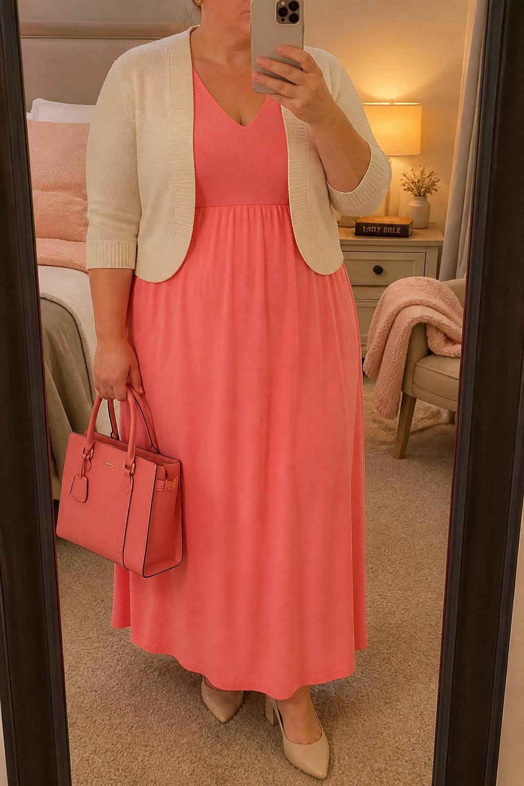 Mirror selfie of a plus size woman wearing a coral pink jersey maxi dress with short sleeves, a modest V-neckline, and a softly fitted empire waist, a cream cropped cardigan worn open, nude pointed block heels, a structured coral handbag, and delicate gold stud earrings, clearly showing the exact clothing items, coral jersey, cream knit, footwear, and accessories listed, holding a phone in front of a mirror, cropped at the neck so the face does not appear, relaxed cheerful selfie pose, in a cozy bedroom mirror setup with evening golden-hour lighting, a folded blanket on a chair and a Bible resting on a bedside table, realistic church outfit, latest iPhone photo quality.