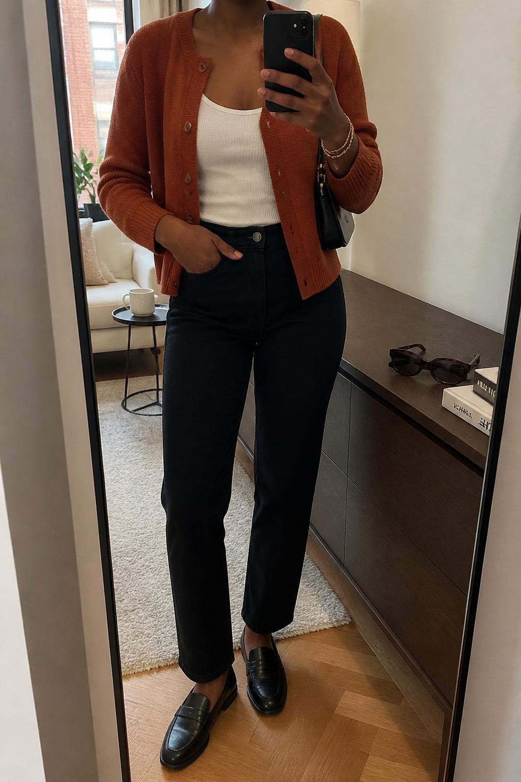 Mirror selfie of a Black woman wearing a rust button-front cardigan sweater, fitted white tank top, black straight-leg jeans, black leather loafers, gold bracelet stack, and a black mini shoulder bag, holding a phone in front of a mirror, cropped at NECK so the face does not show, relaxed casual selfie pose, in a stylish city apartment, soft indoor lighting, with sunglasses placed on a dresser and a coffee mug on a side table, latest iPhone photo quality.