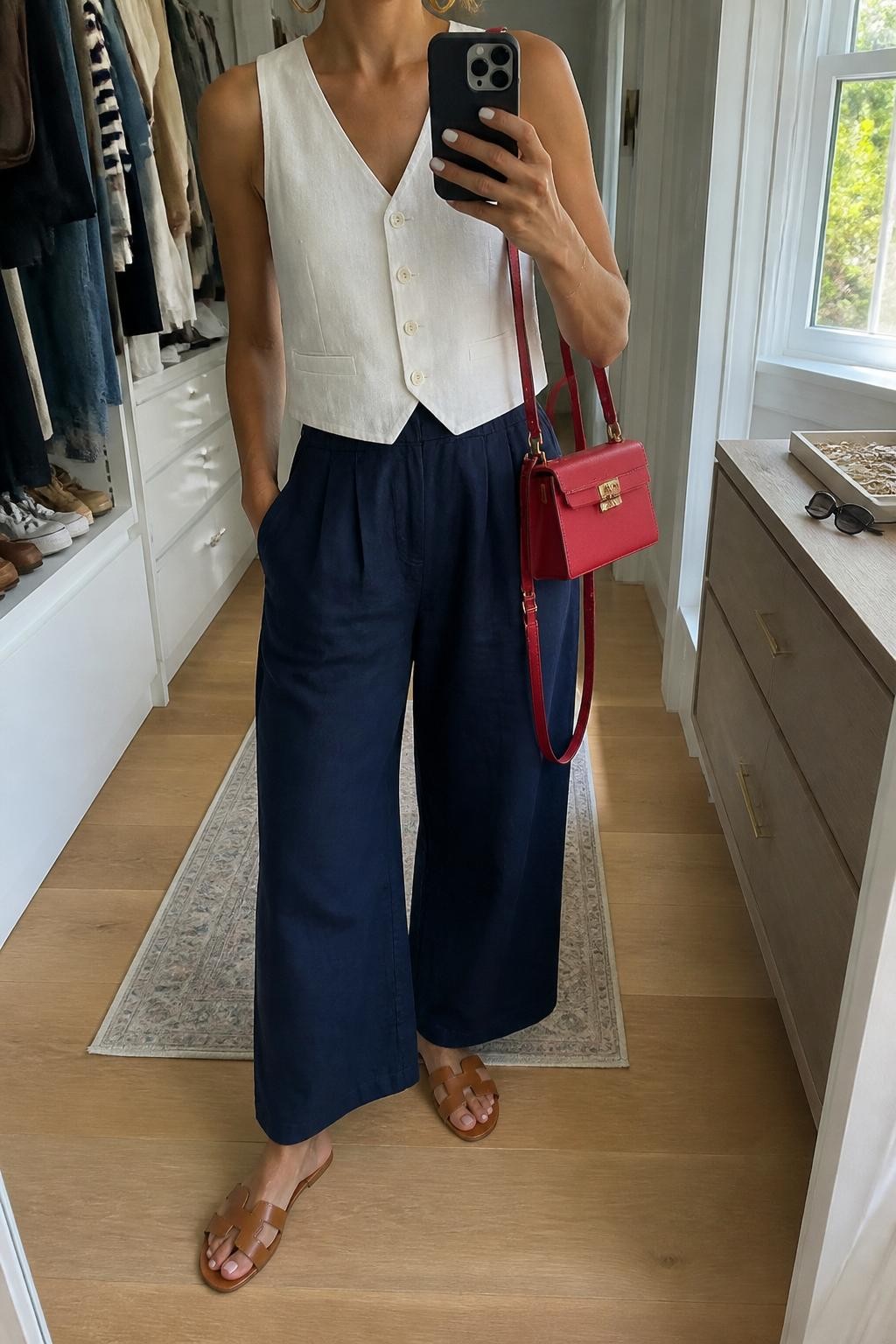 mirror selfie of a woman wearing a fitted white sleeveless linen vest buttoned at the front, navy high-waisted wide-leg linen pants, tan leather slide sandals, red structured mini bag, and gold hoop earrings, clearly showing the same clothing items, colors, fabrics, and accessories listed in the Outfit Pieces section, holding a phone in front of a mirror, cropped at NECK so the face does not show, with a relaxed casual selfie pose, in a walk-in closet mirror, natural window light, including sunglasses placed on a dresser and a jewelry tray on the vanity, latest iPhone photo quality.
