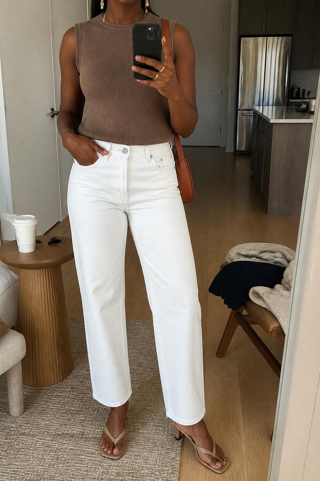 mirror selfie of a Black woman wearing the exact outfit described above, clearly showing a mocha sleeveless fine-knit sweater tucked into white straight-leg jeans, tan kitten-heel sandals, a caramel leather shoulder bag, and gold drop earrings, holding a phone in front of a mirror, cropped at NECK so the face does not show, with a relaxed casual selfie pose, in a bright apartment mirror, natural daylight, with a coffee cup on a side table and folded clothes on a chair, latest iPhone photo quality.