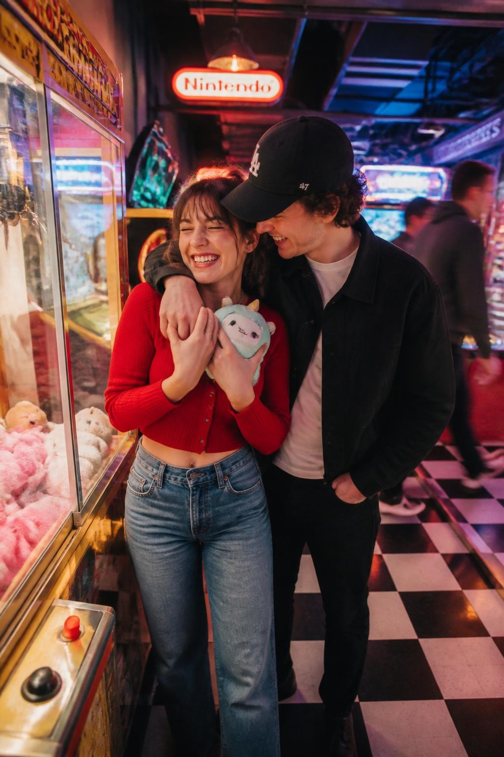 Image prompt: A colorful DSLR couple photo inside a retro arcade, featuring an adult woman with fair skin wearing a red cropped cardigan and high-waisted jeans, holding a small plush prize against her chest while her partner hugs her from the side with one arm around her shoulders, both laughing near a claw machine, neon arcade lights, glowing game screens, coin tray, checkerboard floor, slight motion blur from people in the background, realistic reflections on glass, fun date-night couple photography, candid and joyful.