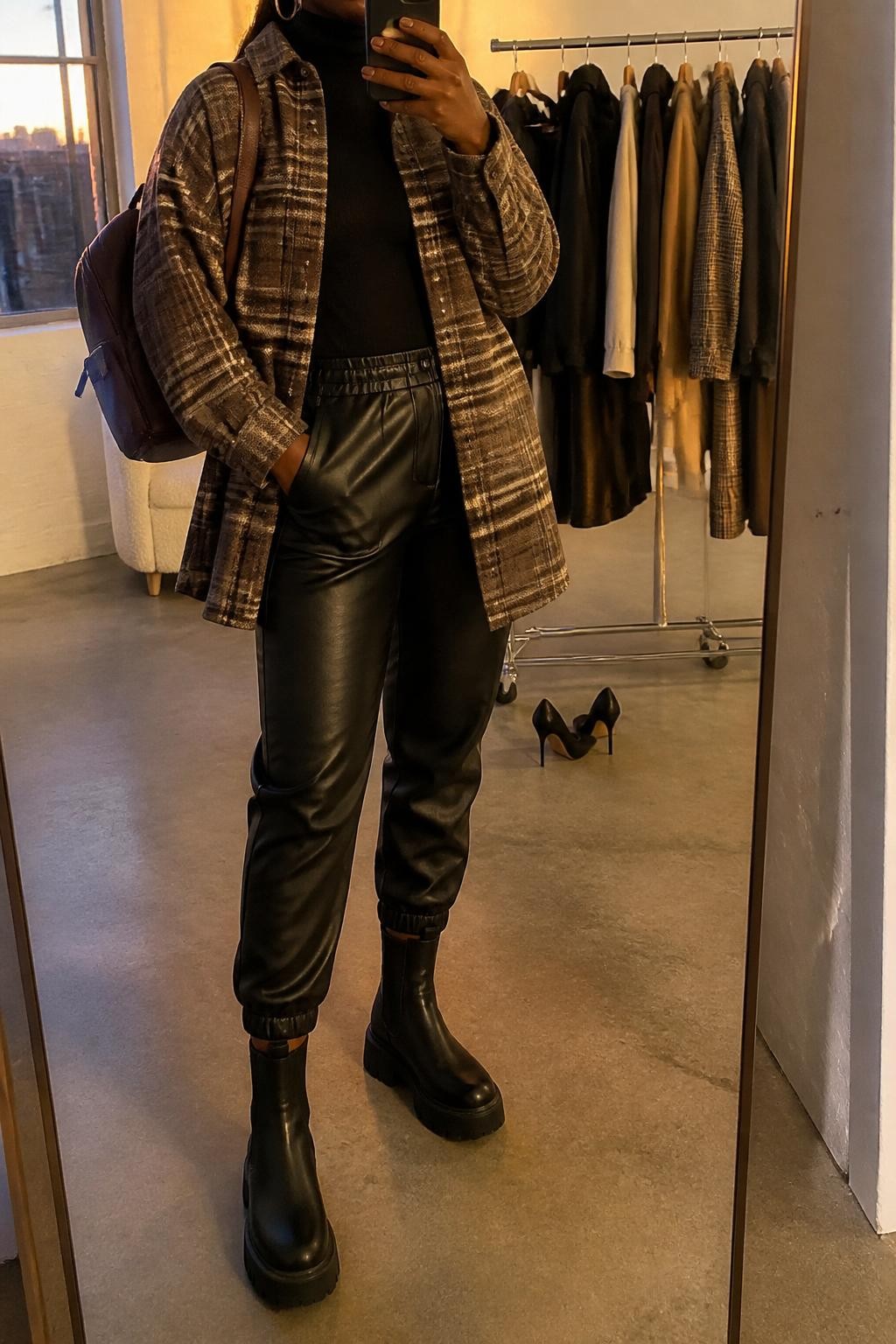 Mirror selfie of a Black woman wearing an oversized brown plaid shacket, fitted black turtleneck, black faux leather joggers, black platform boots, gold hoop earrings, and a dark brown mini backpack, holding a phone in front of a mirror, cropped at NECK so the face does not show, relaxed casual selfie pose, in a fashion studio mirror corner, evening golden hour, with a clothing rack with garments behind and a pair of heels on the floor nearby, latest iPhone photo quality.