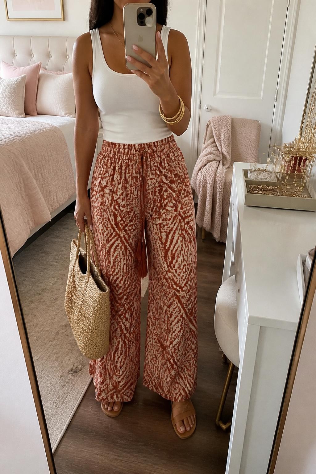 Mirror selfie of a Latina woman wearing a fitted white tank top, rust and cream printed wide-leg pants, tan flat sandals, gold bangles, and a straw tote bag, holding a phone in front of a mirror, cropped at NECK so the face does not show, relaxed casual selfie pose, in a feminine-themed bedroom, soft indoor lighting, with a folded blanket on a chair and a jewelry tray on the vanity, latest iPhone photo quality.