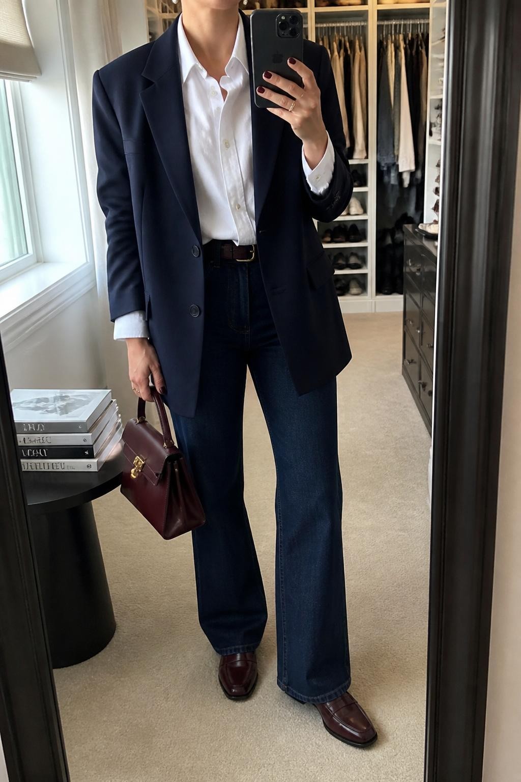 mirror selfie of a woman wearing the exact outfit described above, clearly showing indigo trouser jeans, a crisp white button-down shirt, a navy oversized blazer, burgundy loafers, a burgundy top-handle bag, and pearl stud earrings, holding a phone in front of a mirror, cropped at NECK so the face does not show, with a relaxed casual selfie pose, in a walk-in closet mirror, natural window light, with an open closet with clothes visible and a stack of fashion magazines on a side table, latest iPhone photo quality.