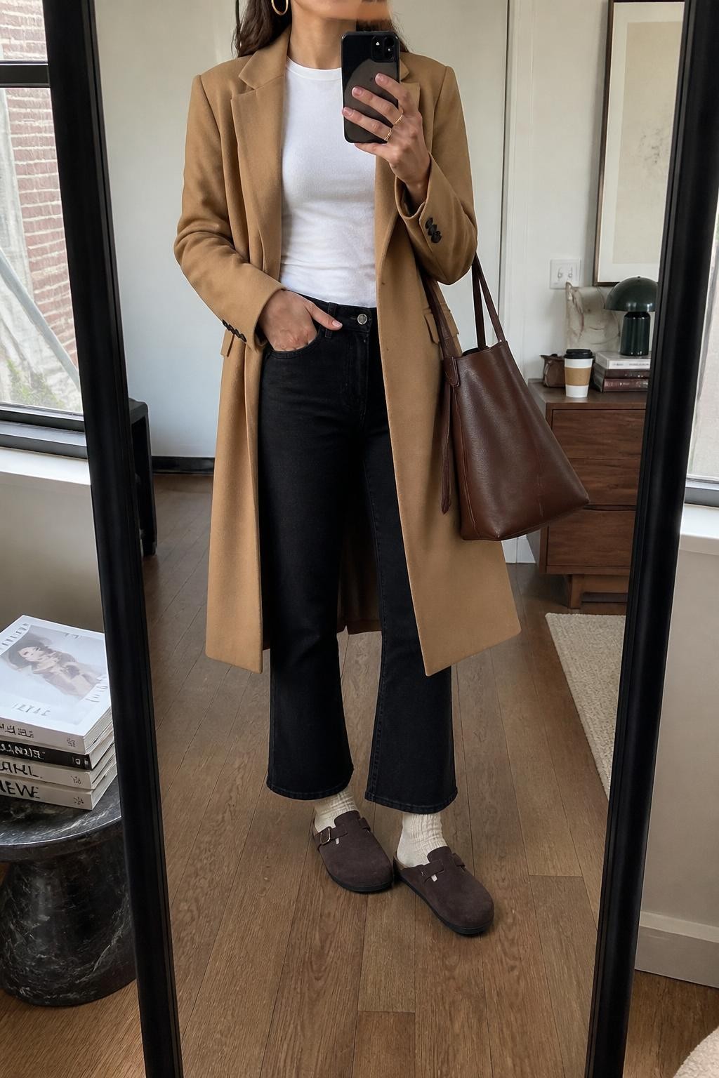 mirror selfie of a woman wearing the exact outfit described above, clearly showing brown buckle Birkenstock-style clogs with cream socks, black cropped flare jeans, a fitted white tee, a camel long coat worn open, a brown leather tote bag, and gold hoop earrings, holding a phone in front of a mirror, cropped at NECK so the face does not show, with a relaxed casual selfie pose, in a stylish city apartment, natural window light, with a stack of fashion magazines on a side table and a coffee cup on a dresser, latest iPhone photo quality.