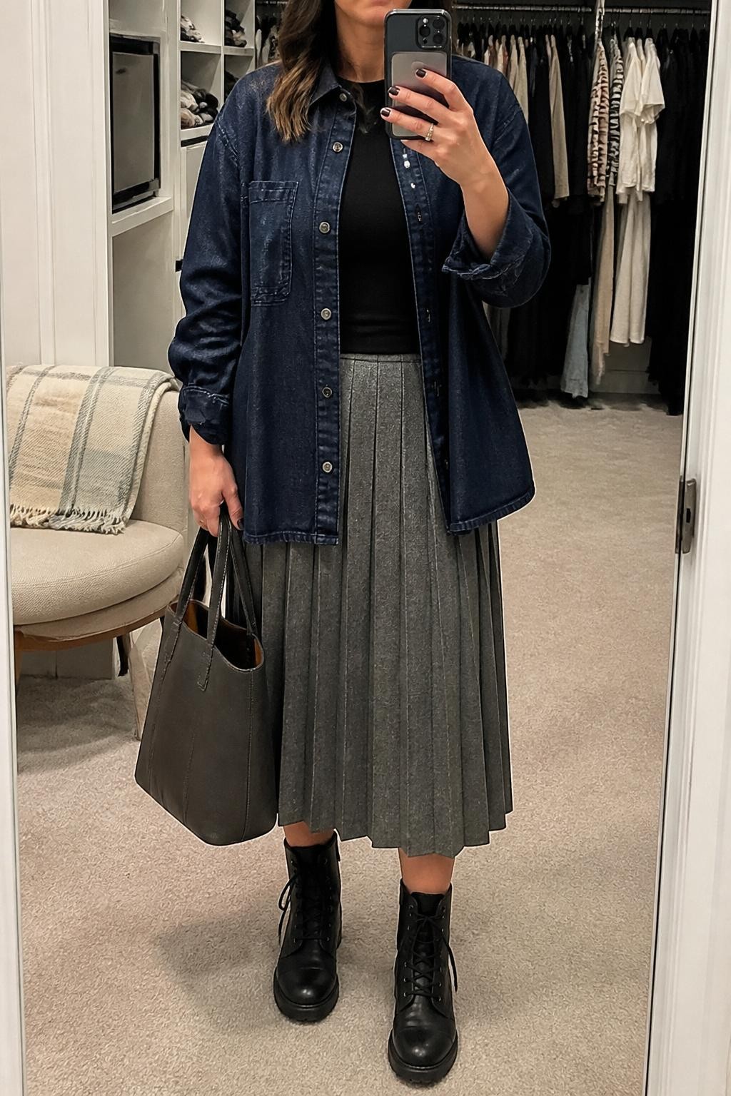 Mirror selfie of a woman wearing a dark indigo denim overshirt over a fitted black crew-neck sweater and a heather gray pleated midi skirt, clearly showing black lace-up ankle boots, a dark gray leather tote, and small silver stud earrings, holding an iPhone in front of a walk-in closet mirror, cropped at the neck so the face does not show, relaxed casual selfie pose, soft indoor lighting, an open closet with clothes visible and a folded blanket on a chair, natural unstaged setting, latest iPhone photo quality.