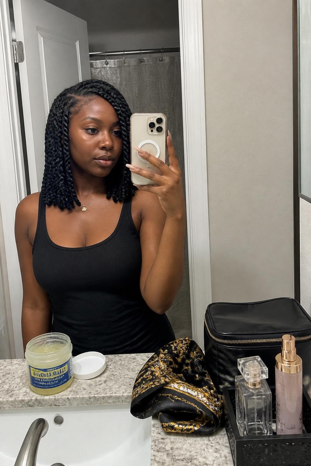 Image prompt: Photo of a Black woman wearing a shoulder-length Senegalese twist bob with a deep side part and smooth rope texture, candid mirror selfie, bathroom vanity mirror with perfume and a makeup bag nearby, soft indoor lighting, containing an open jar of braiding gel on the counter and a silk scarf beside the sink, realistic morning moment, latest iPhone photo quality.