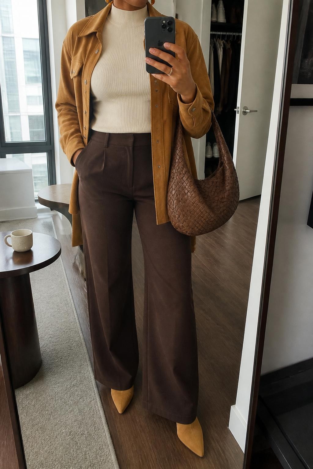 Mirror selfie of a Black woman wearing a caramel suede shacket over a fitted cream mock-neck top, clearly showing dark chocolate wide-leg trousers, pointed tan ankle boots, a woven brown handbag, and small amber earrings, holding an iPhone in front of a stylish city apartment mirror, cropped at the neck so the face does not show, relaxed casual selfie pose, natural window light, a coffee mug on a side table and an open closet with clothes visible, natural unstaged setting, latest iPhone photo quality.