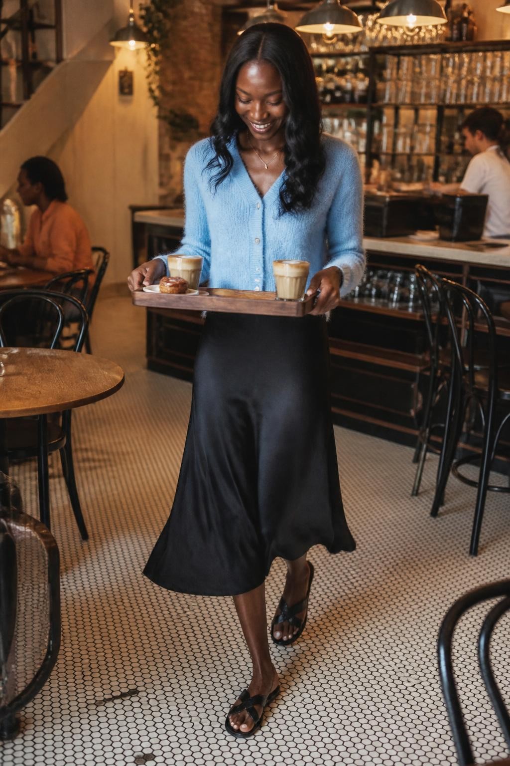 A realistic DSLR cafe photo of an adult Black woman with deep brown skin walking carefully with a wooden tray holding two lattes and a small pastry, wearing a soft blue cardigan, black slip skirt, and flat sandals, smiling down at the tray, elbows slightly bent, shoulders relaxed, cafe tables and chairs around her, warm pendant lights, tiled floor, bar counter in the background, realistic coffee foam, slight motion in her skirt, natural expression, stylish brunch cafe mood, human candid photography.