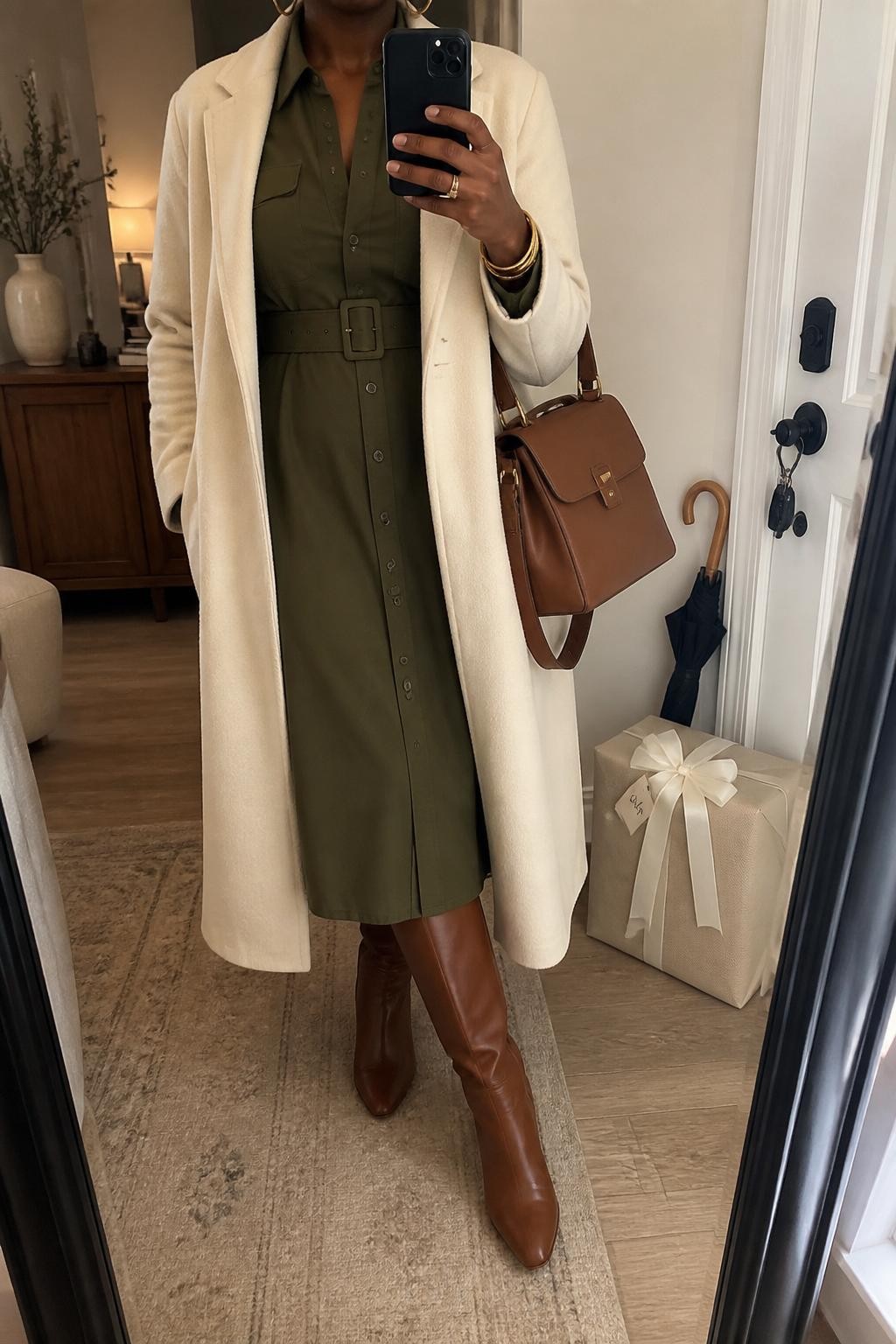 mirror selfie of a Black woman wearing the exact outfit described above, clearly showing an olive green belted shirt midi dress, cognac leather knee-high boots, a cream wool coat worn open, a brown structured satchel bag, and gold hoop earrings, holding a phone in front of a mirror, cropped at NECK so the face does not show, with a relaxed casual selfie pose, in a cozy apartment entry mirror, soft indoor lighting, with a wrapped neutral baby shower gift and umbrella near the door, latest iPhone photo quality.