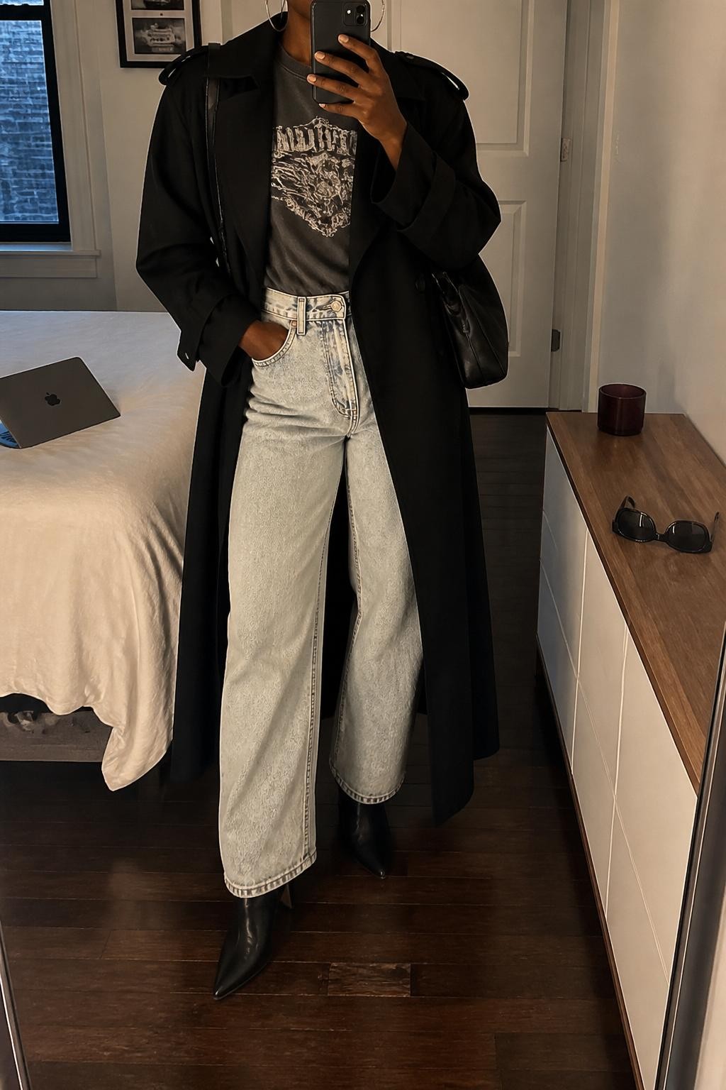 Mirror selfie of a Black woman wearing a black long trench coat, vintage-style gray graphic tee, light wash baggy jeans, black pointed ankle boots, silver hoop earrings, and a black shoulder bag, holding a phone in front of a mirror, cropped at NECK so the face does not show, relaxed casual selfie pose, in a stylish city apartment, warm moody lighting, with a laptop on the bed and sunglasses placed on a dresser, latest iPhone photo quality.