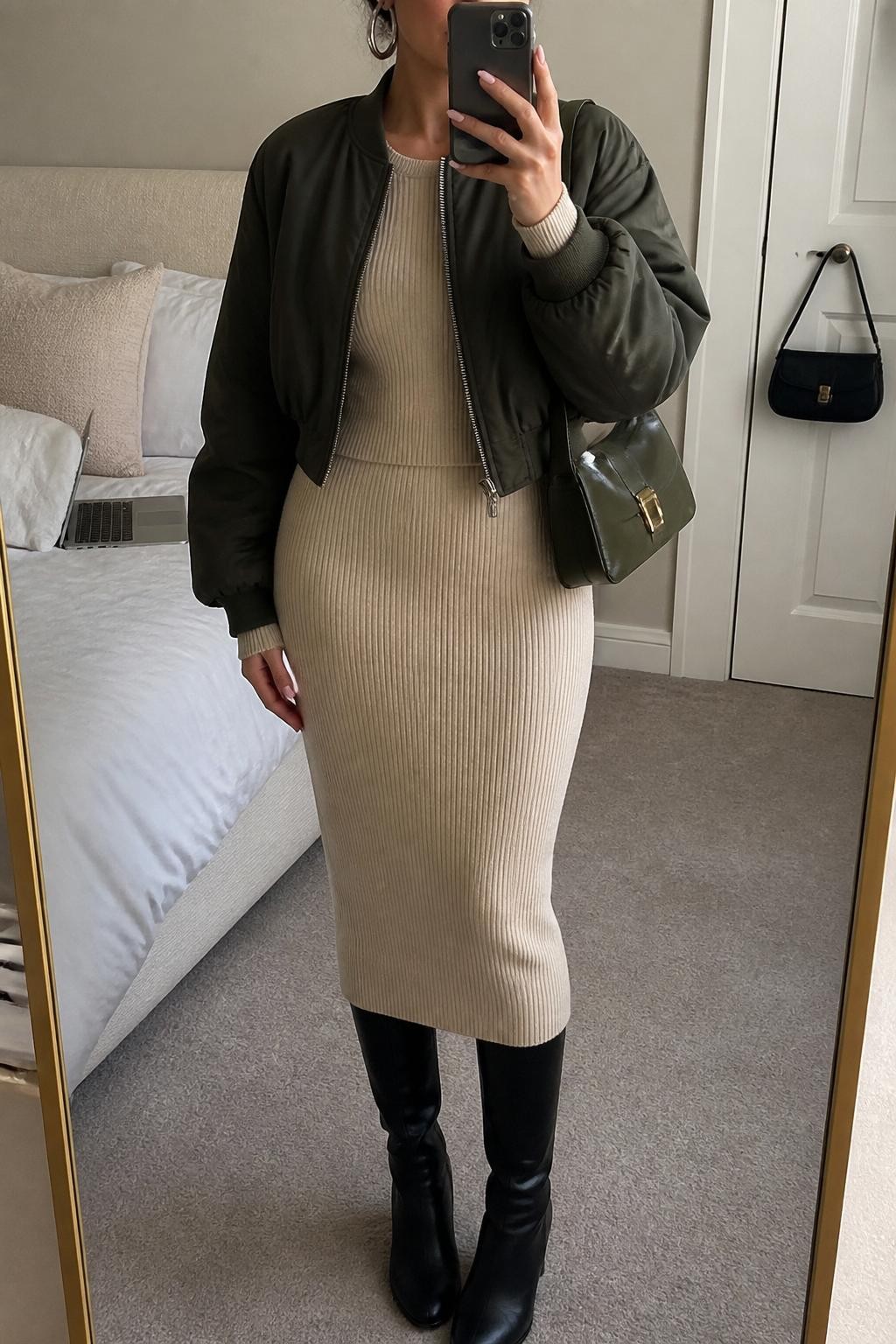 Mirror selfie of a woman wearing a dark olive cropped bomber jacket over a fitted beige ribbed long-sleeve top with a matching beige ribbed midi skirt, clearly showing black knee-high boots, a structured olive shoulder bag, and chunky silver hoop earrings, holding an iPhone in front of a cozy bedroom mirror setup, cropped at the neck so the face does not show, relaxed casual selfie pose, soft indoor lighting, a laptop on the bed and a handbag hanging on a door handle, natural unstaged setting, latest iPhone photo quality.